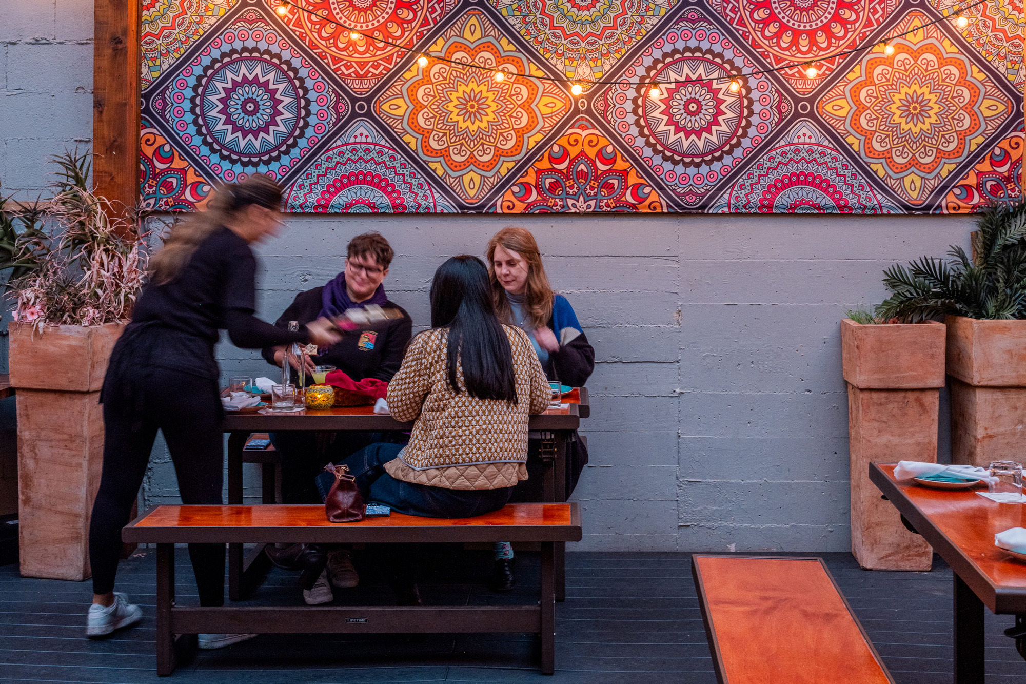 Four people sit at a wooden table in a restaurant with a vibrant, patterned wall tapestry.