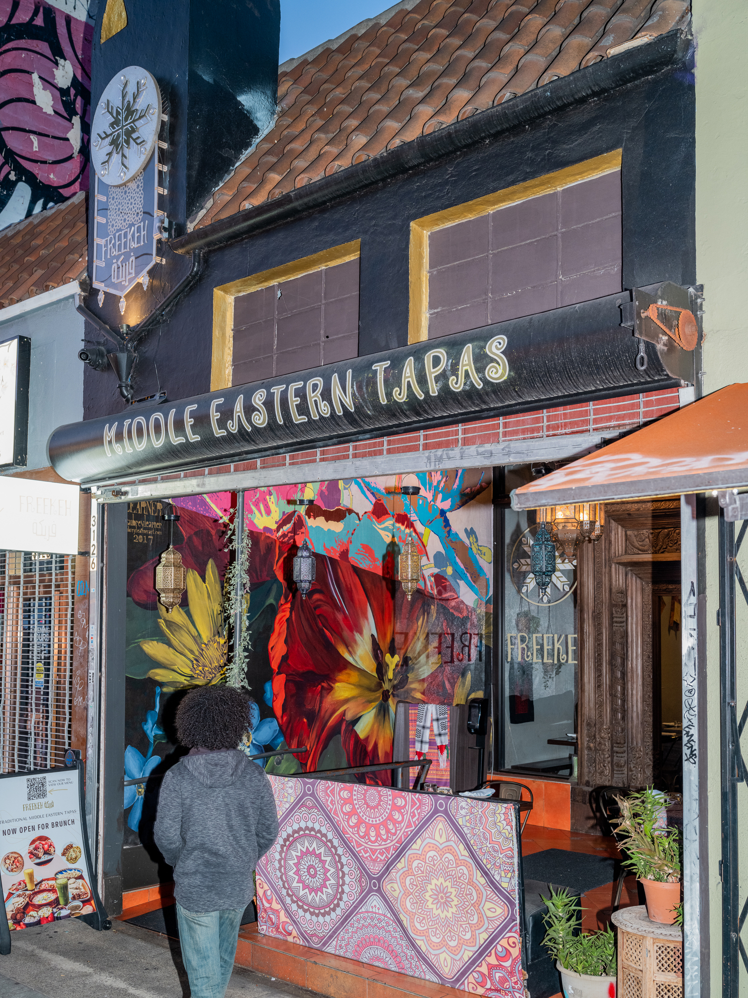 The image shows a vibrant Middle Eastern tapas restaurant with floral wall art, decorative lanterns, and a person in a hoodie approaching the entrance.