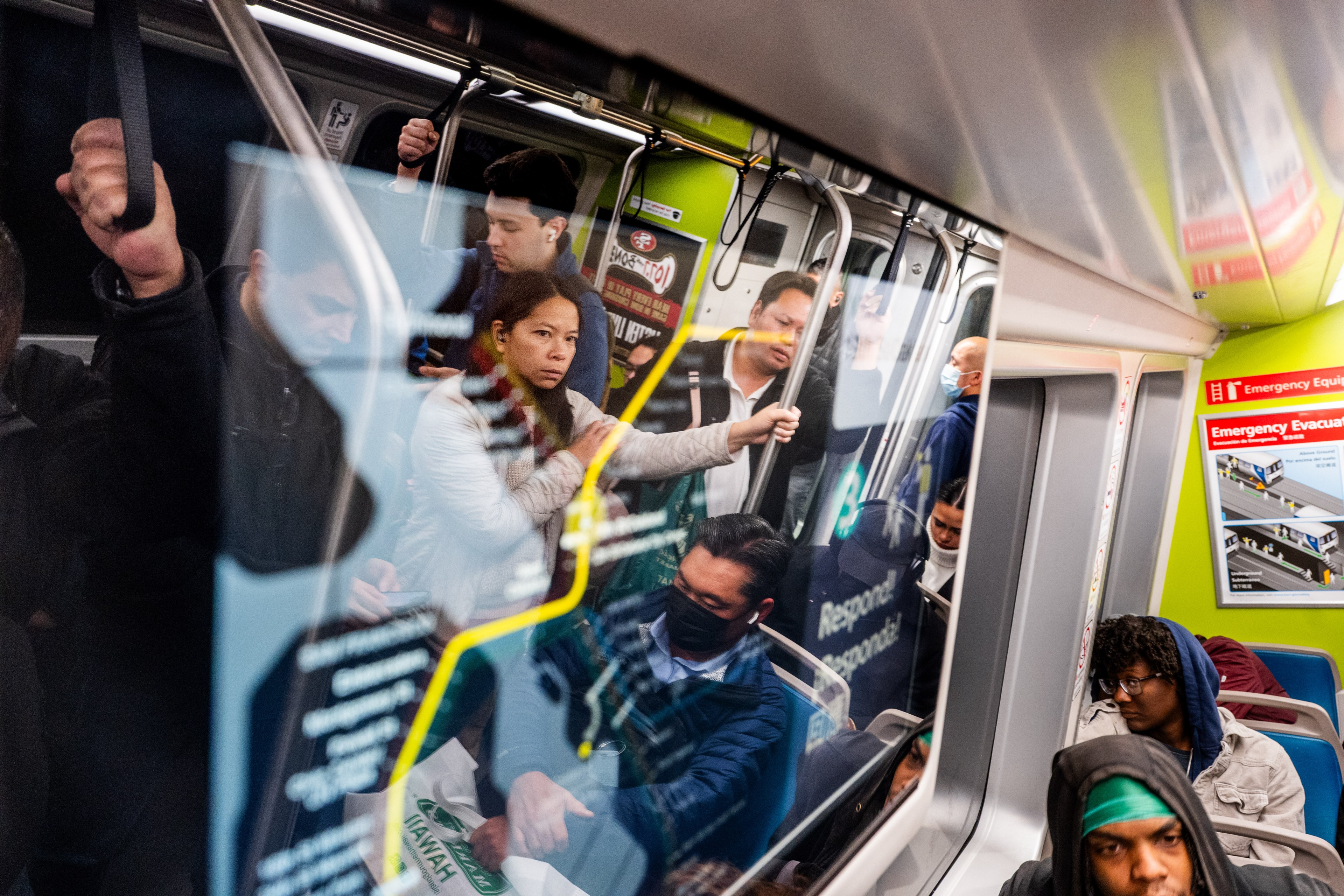 A group of people stands and sits inside a crowded subway car. Some are holding onto straps, and others are wearing masks. Reflections are visible on the window.