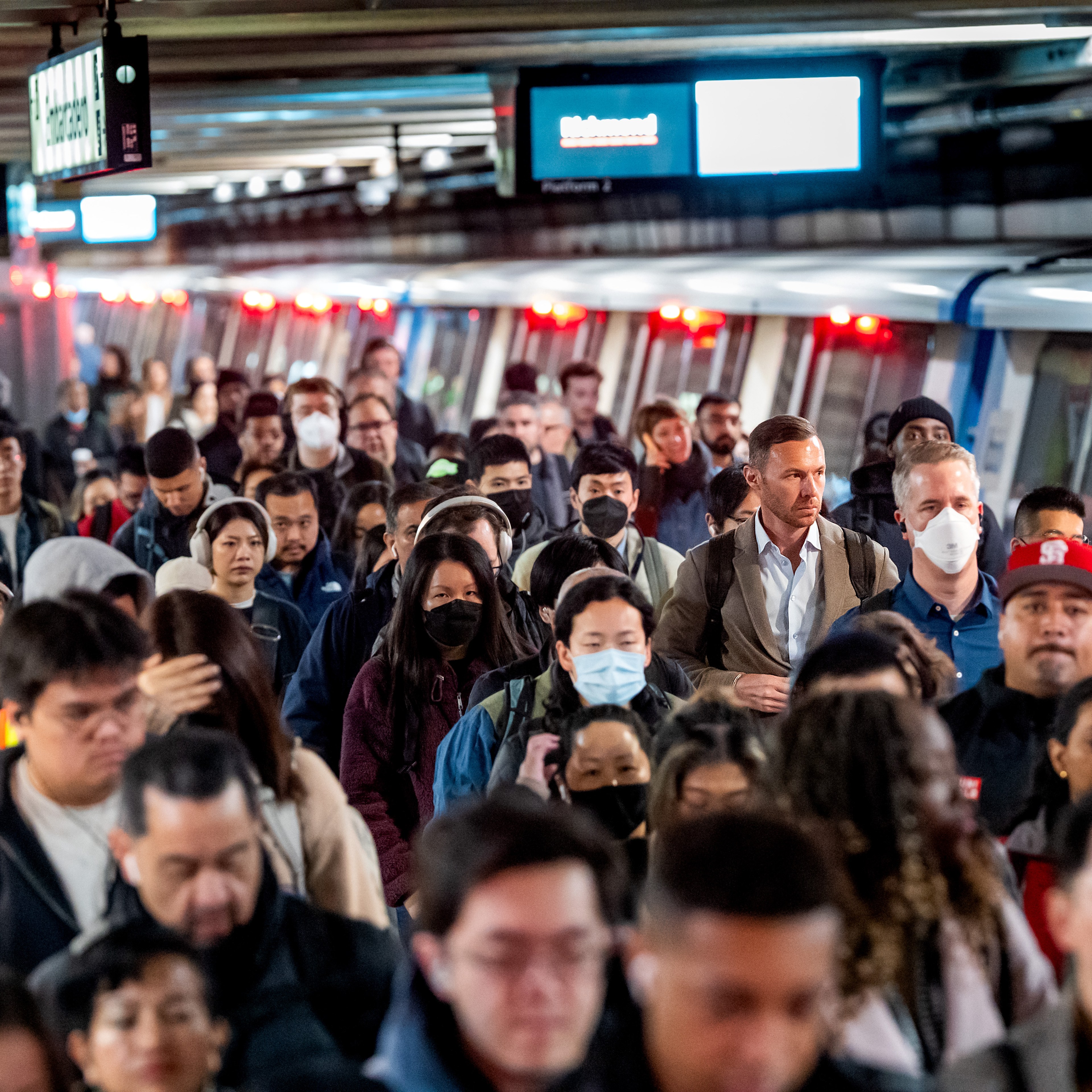 A crowded train platform with diverse people, some wearing masks, are exiting and moving in different directions. The train is visible on the right.