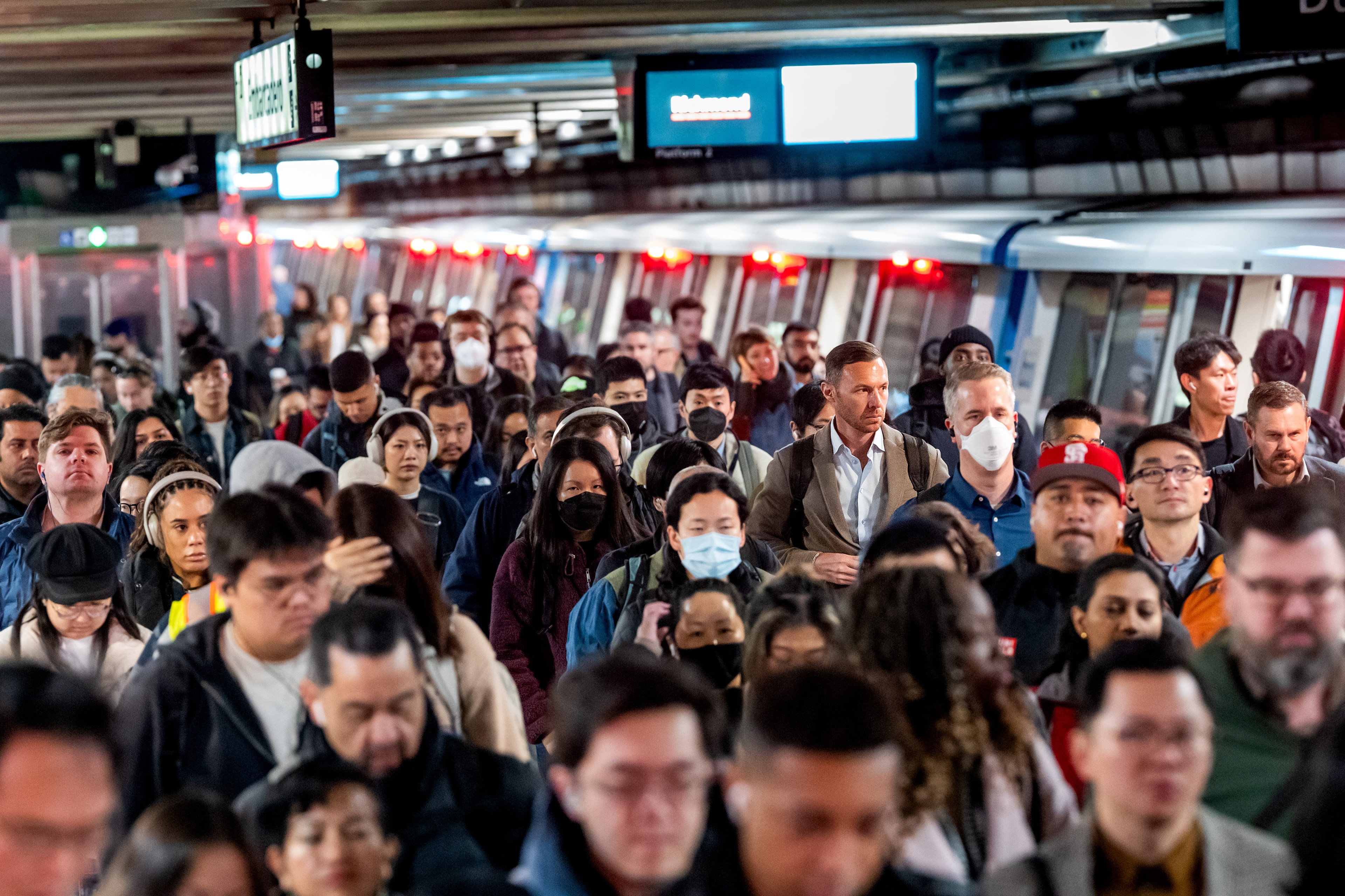 A crowded train platform with diverse people, some wearing masks, are exiting and moving in different directions. The train is visible on the right.