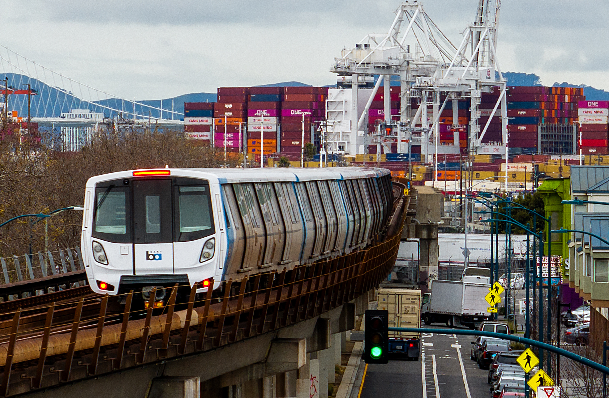 A train travels on an elevated track with a busy shipping port in the background. Shipping containers and cranes are visible under a cloudy sky.