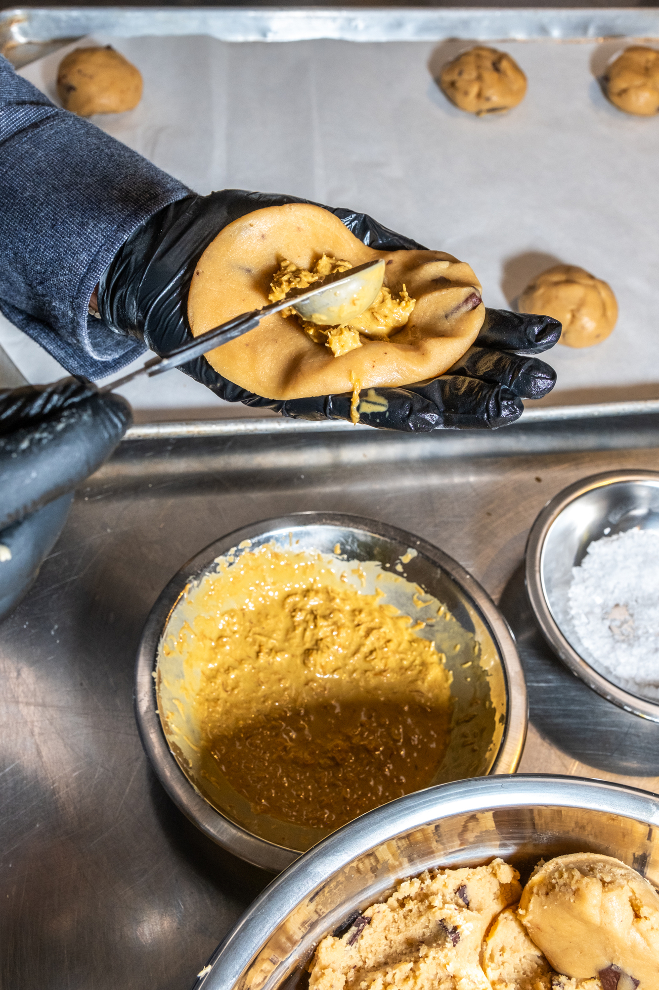 A gloved hand holds and fills cookie dough with a spoonful of a yellow mixture. Nearby bowls hold the mixture, dough, and salt, with cookies on a sheet.