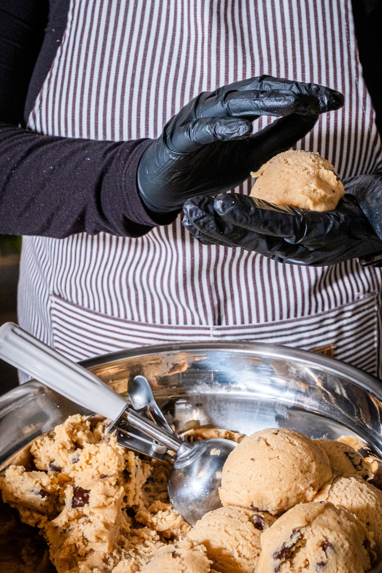 A person wearing black gloves and a striped apron shapes cookie dough over a metal bowl filled with more dough and a scooper.