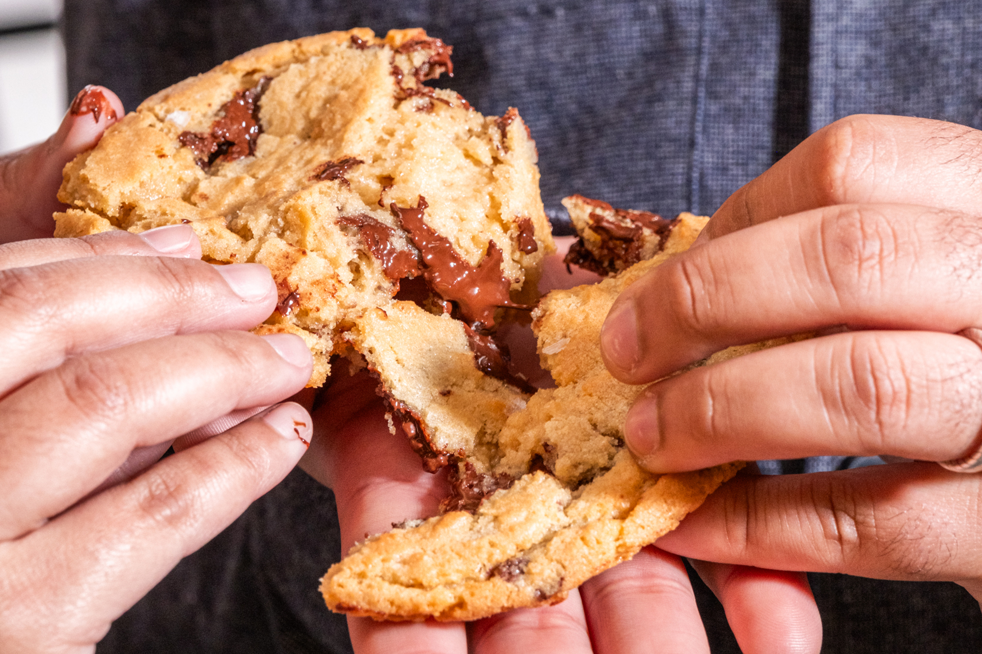 Two hands pull apart a large, gooey chocolate chip cookie, revealing melted chocolate chunks inside. The background is a blurred dark fabric.