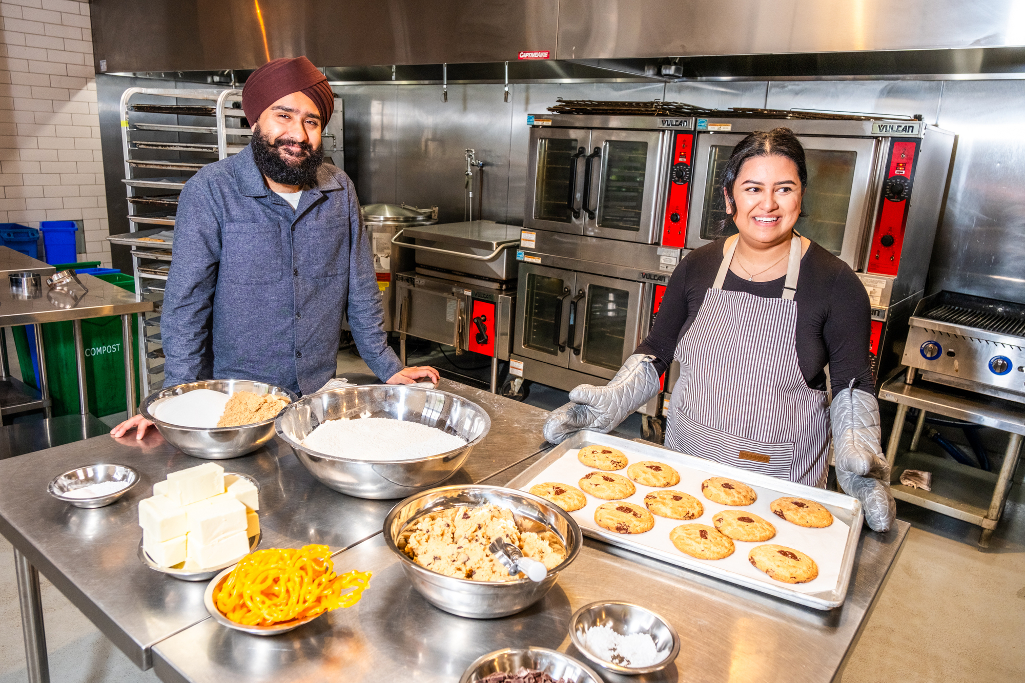 Two people in a professional kitchen stand near a table with baking ingredients. One holds a tray of cookies, and ovens are visible in the background.