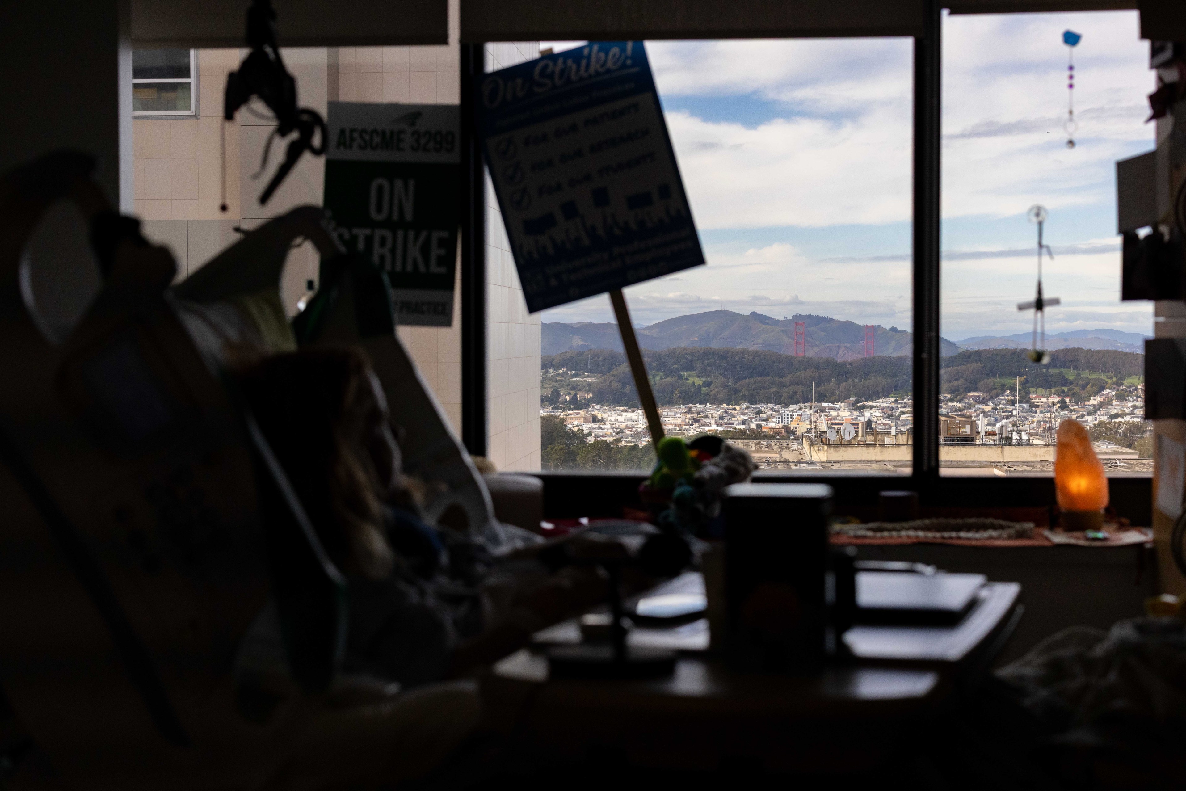 A hospital room with a bed and 'On Strike' signs. Through the window, mountains, the Golden Gate Bridge, and a cityscape are visible.