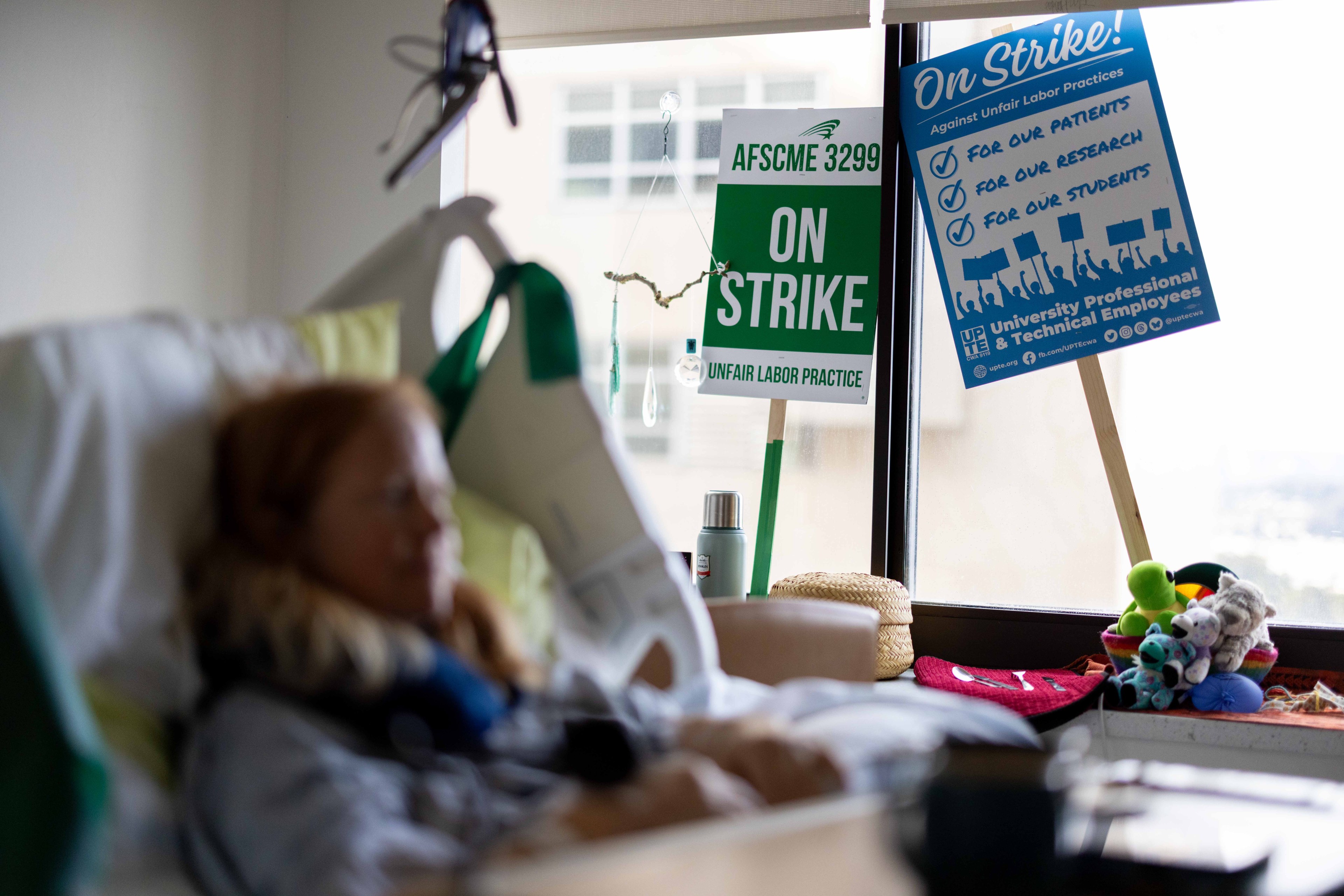 A person in a hospital bed lies unfocused in the foreground. In the background, there are two strike signs in a window with toys and a thermos nearby.