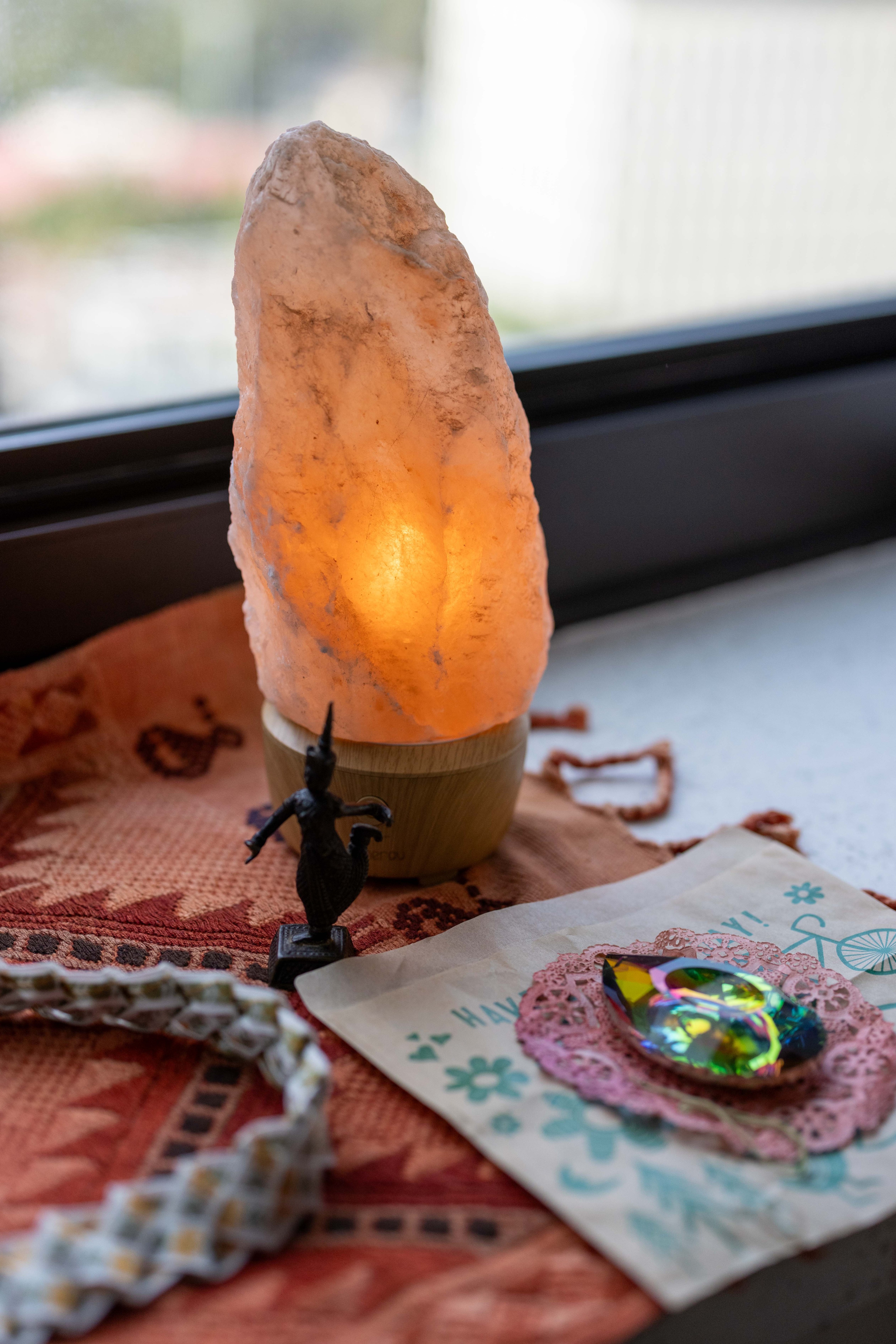A glowing salt lamp sits on a patterned cloth next to a small figurine. Nearby, there's a colorful, reflective crystal on a pink doily atop paper.
