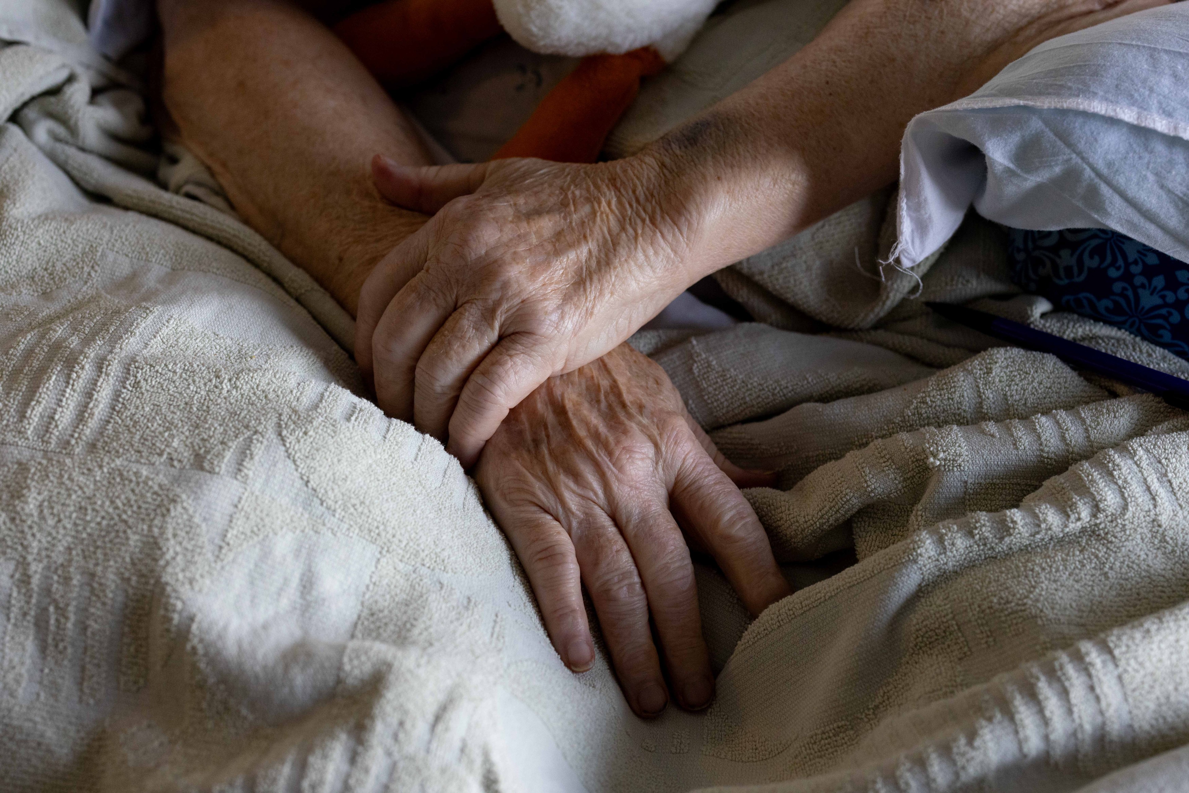 Two elderly hands rest gently atop each other on a textured beige blanket, conveying a sense of calm and tenderness.