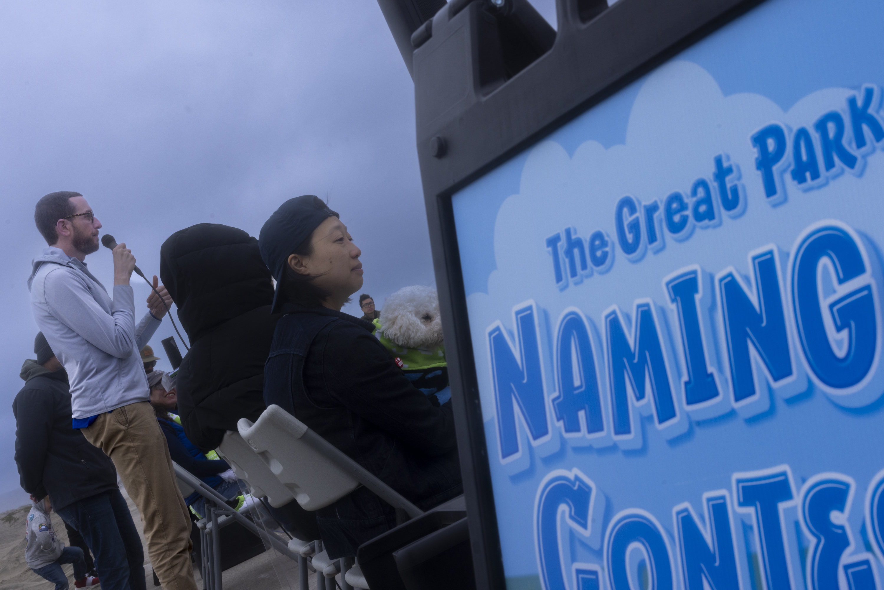 A man is speaking into a microphone at an outdoor event. People sit nearby, and a sign reads, &quot;The Great Park Naming Contest.&quot;