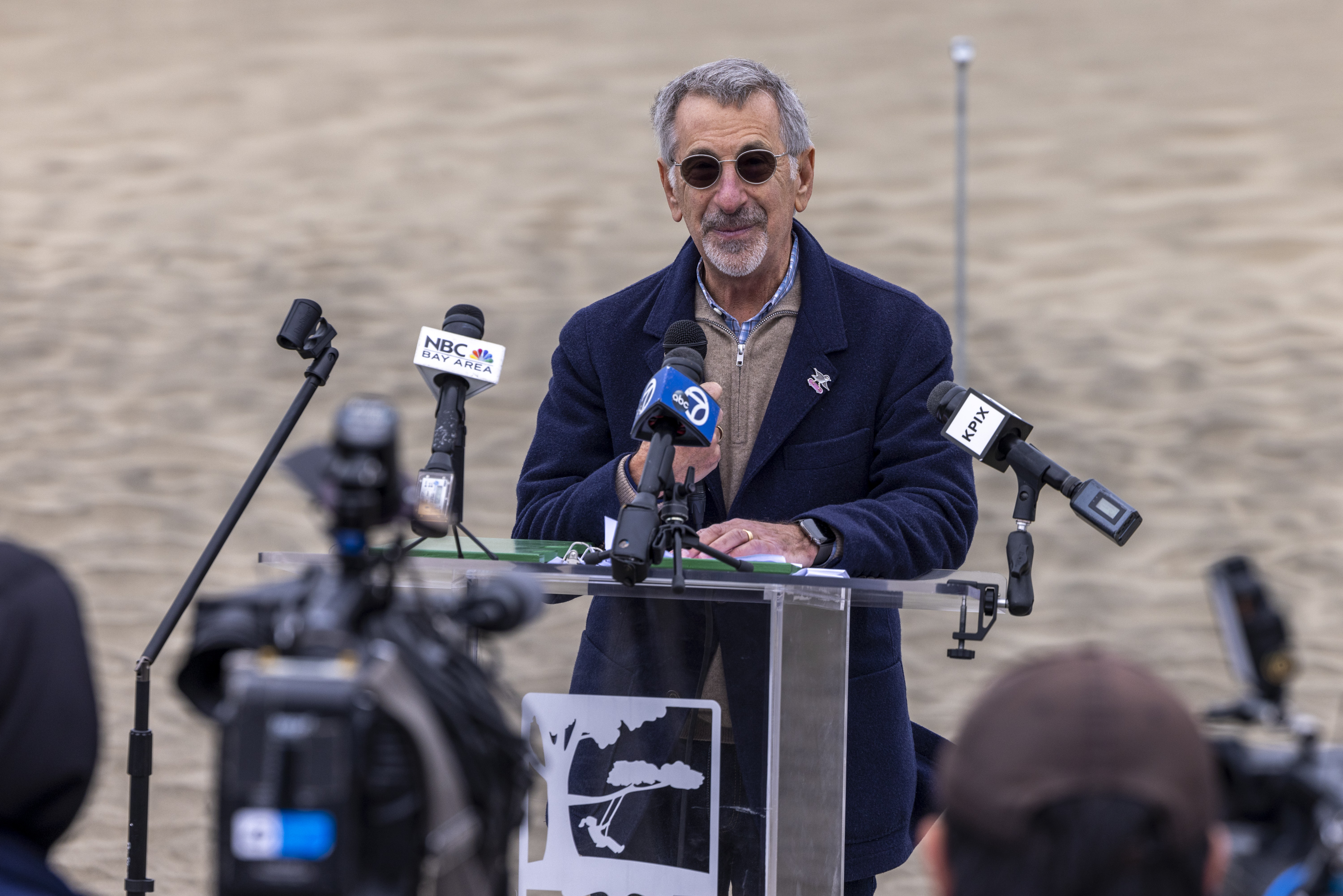 A man in glasses speaks at a podium outdoors, surrounded by microphones labeled with various media outlets like NBC Bay Area and KPIX.