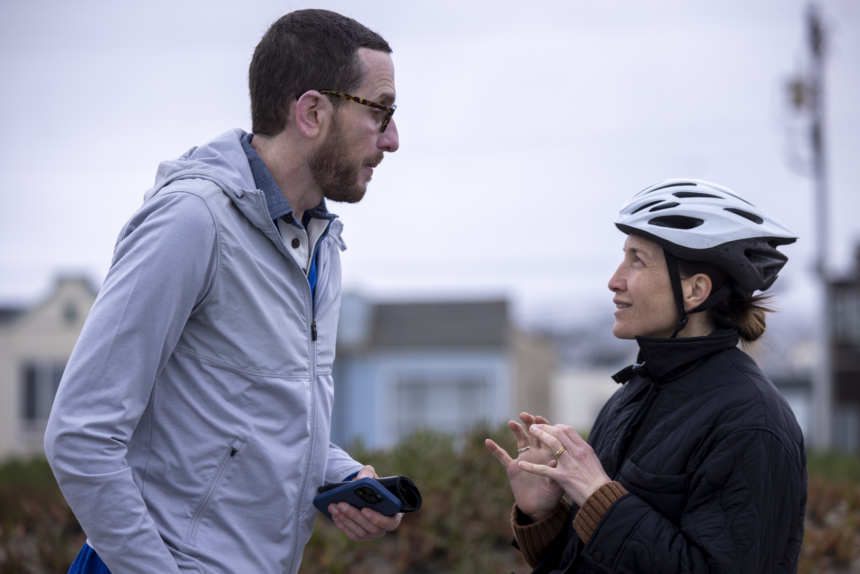 A man in a gray jacket and glasses holds a phone while talking to a woman wearing a black jacket and a white bike helmet, with houses blurred in the background.