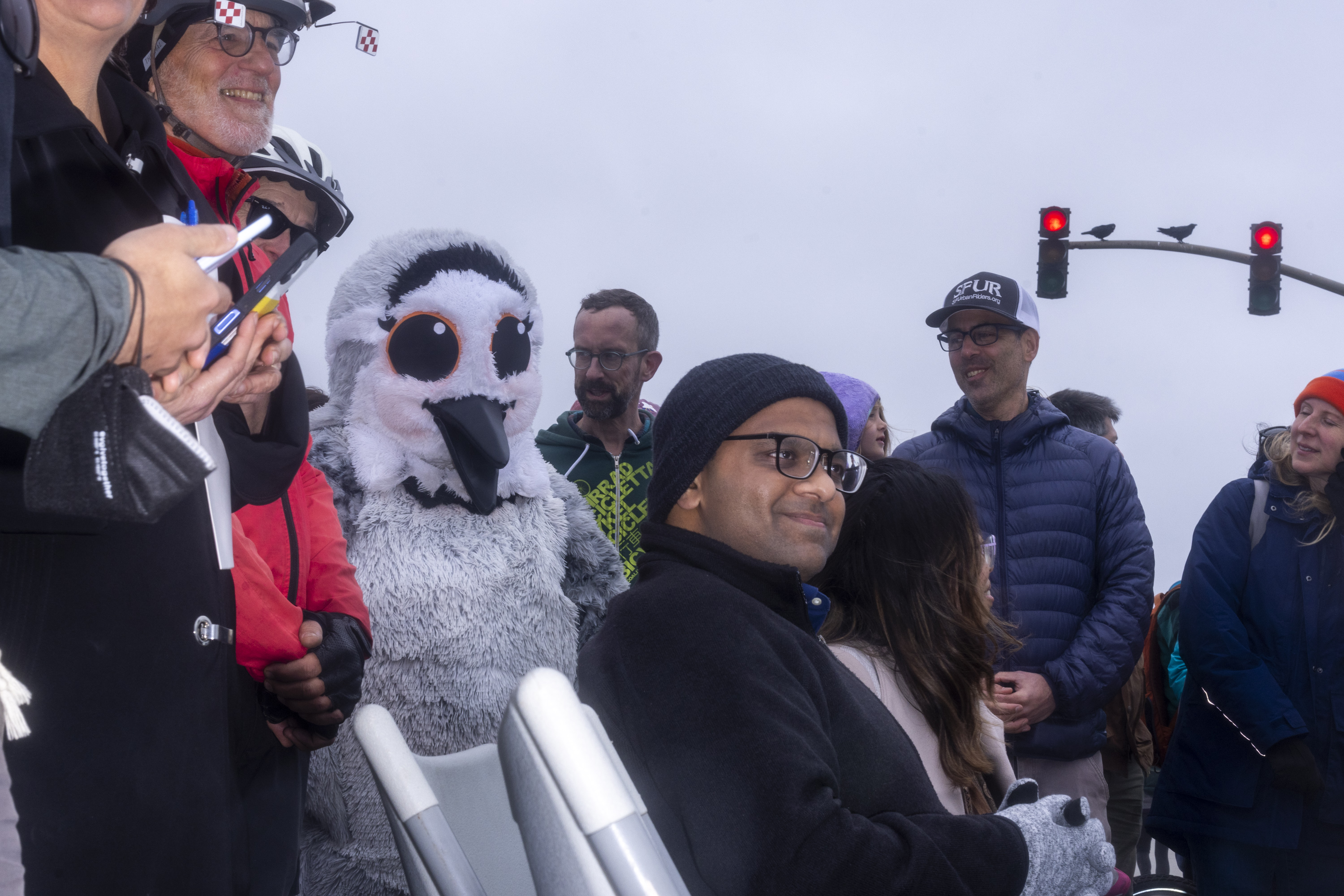 A group of people stands together, including someone in a fluffy bird costume with large eyes. They're outdoors, near a traffic light.