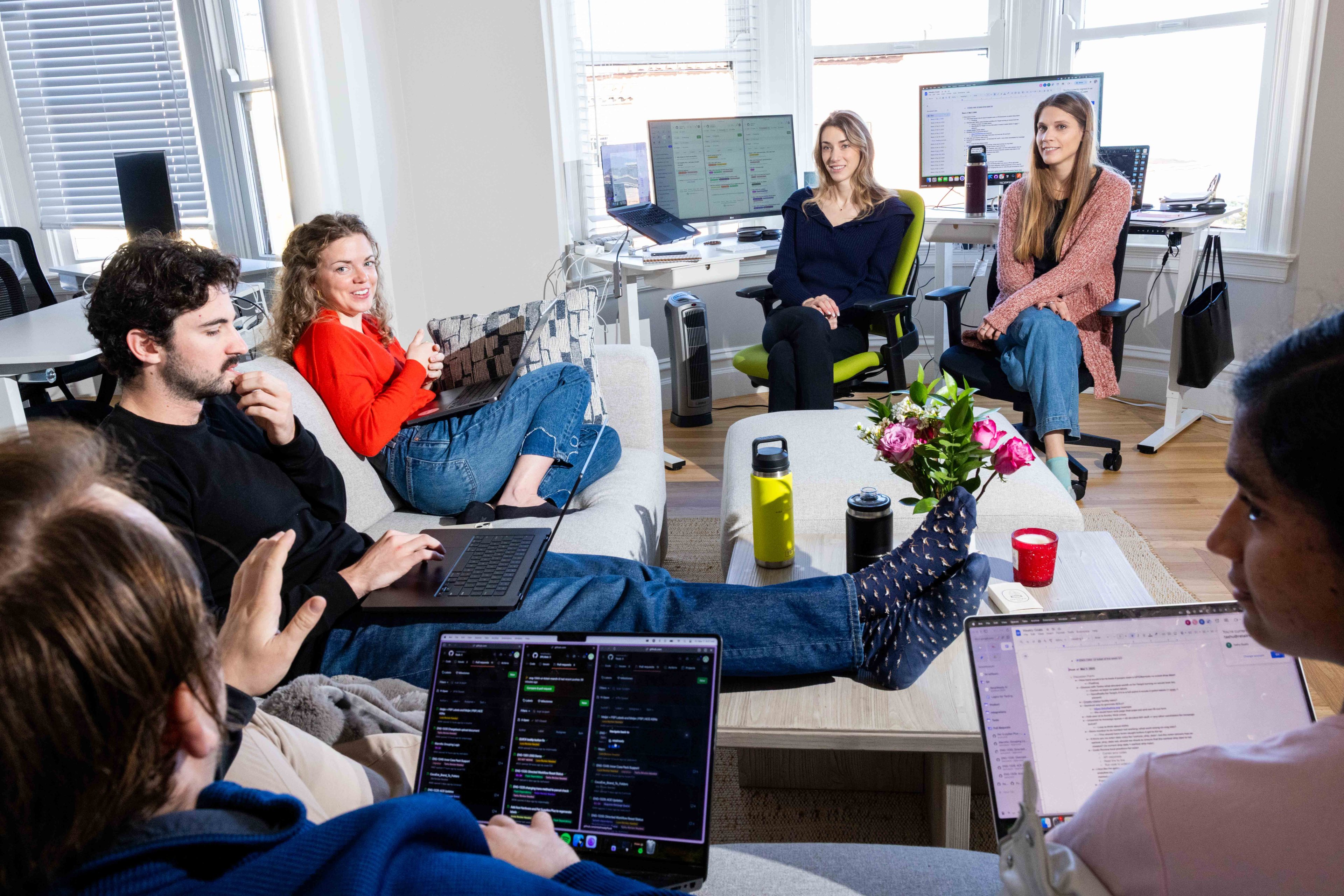 A group of six people sit in a bright room with laptops, casually interacting. Some are on a sofa, others in chairs. Monitors and flower vases are on tables nearby.