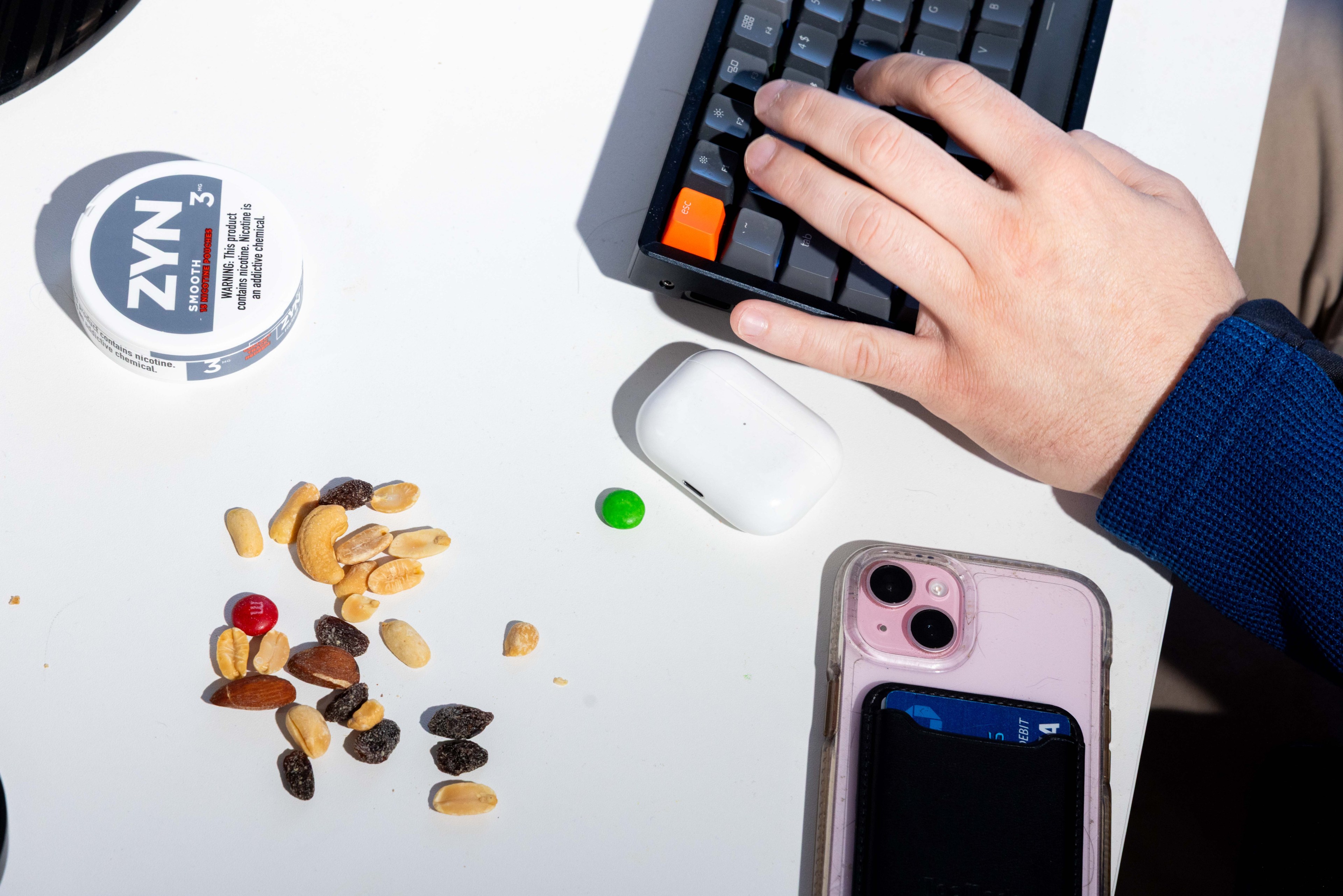 A hand uses a keyboard beside an open tin of ZYN, a mix of nuts and candies, AirPods case, and a pink smartphone with a card holder on a white surface.