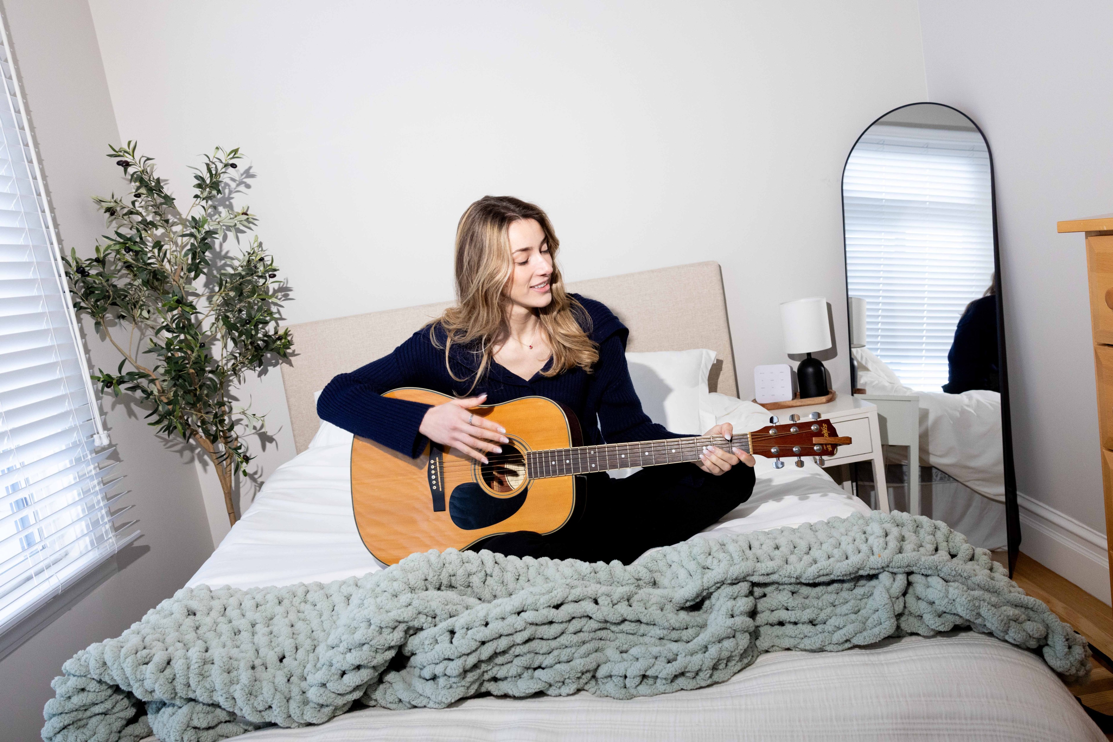 A person sits on a bed playing an acoustic guitar. The room has a potted plant, a mirror, and a bedside table with a lamp. A knitted blanket covers the bed.