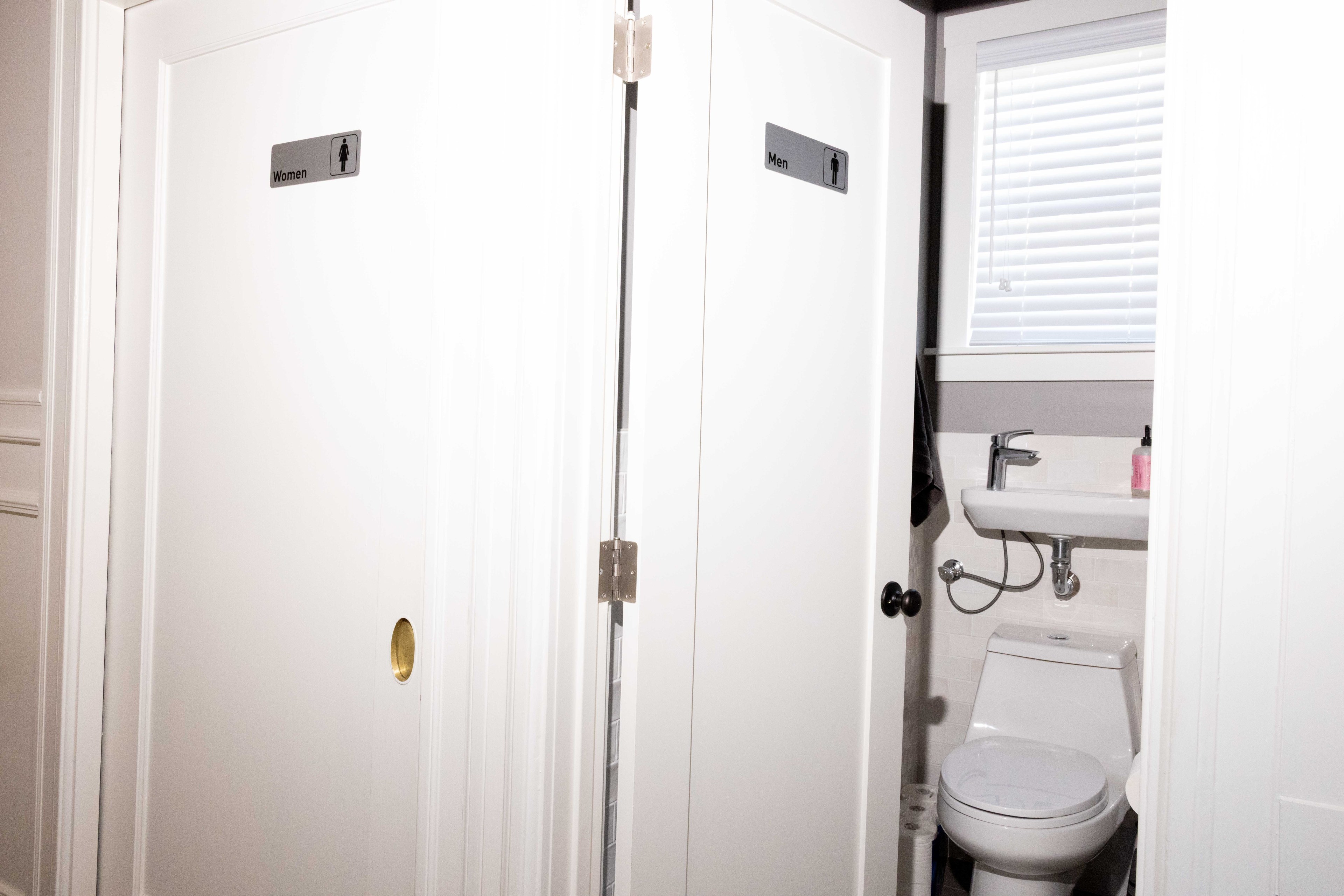 The image shows two restroom doors with signs for &quot;Women&quot; and &quot;Men.&quot; The men's door is slightly open, revealing a small bathroom with a toilet and sink.