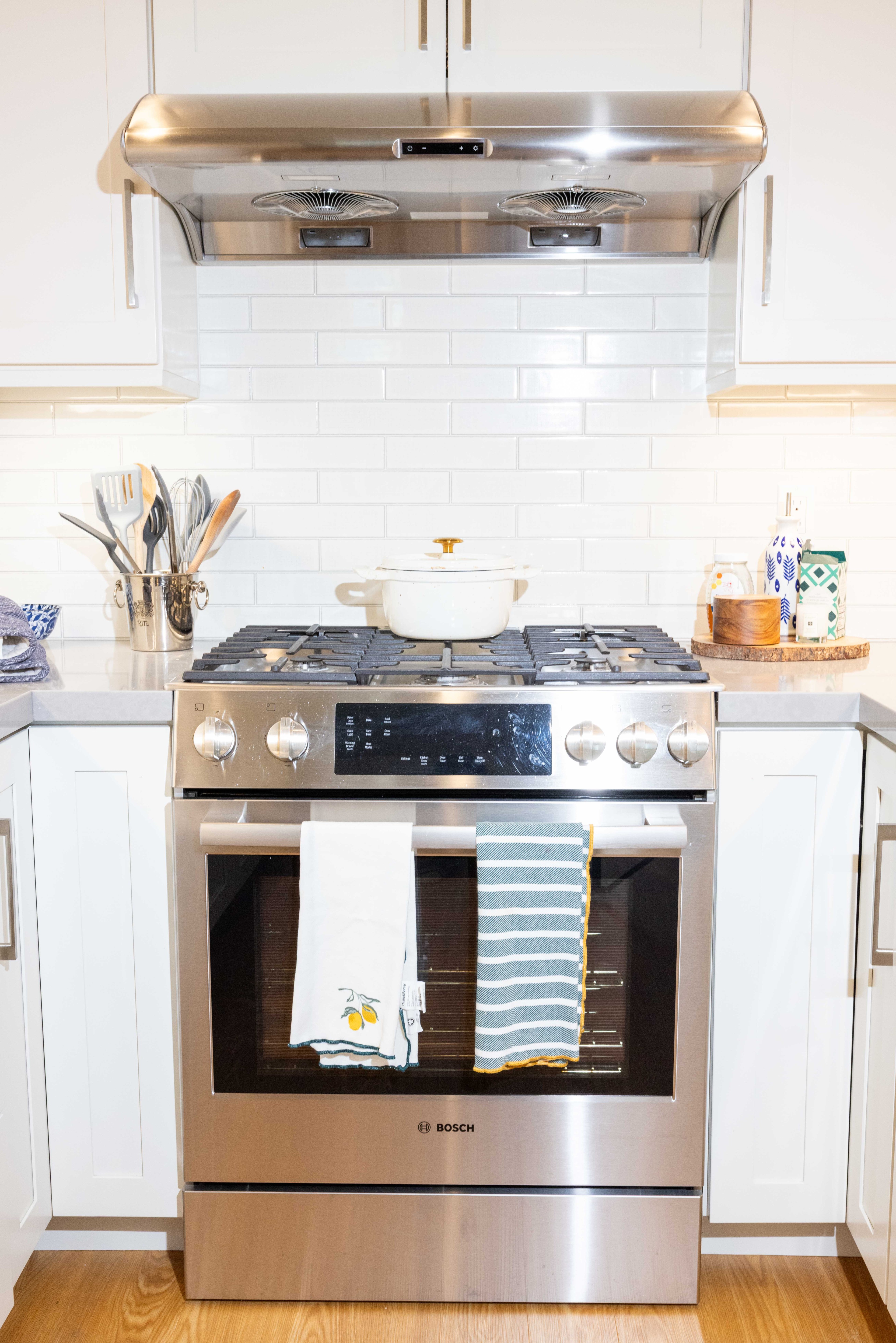 A modern kitchen stove with a white pot sits under a stainless steel range hood. Two towels hang from the oven door, and utensils are in a holder nearby.