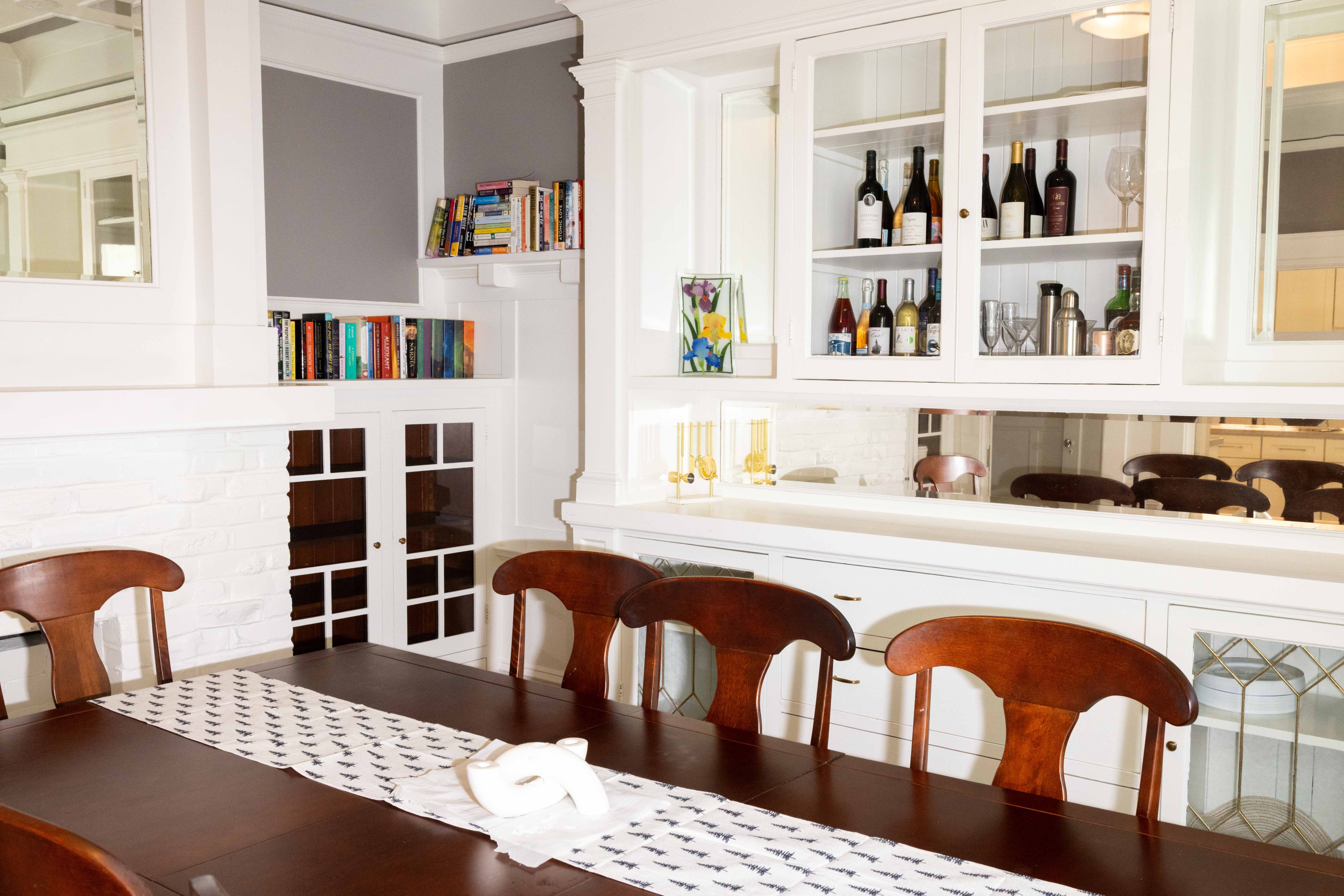 The image shows a dining room with a dark wooden table and chairs, a white runner, and decorative ceramics. There's a cabinet with wine bottles and glasses, plus books on shelves.