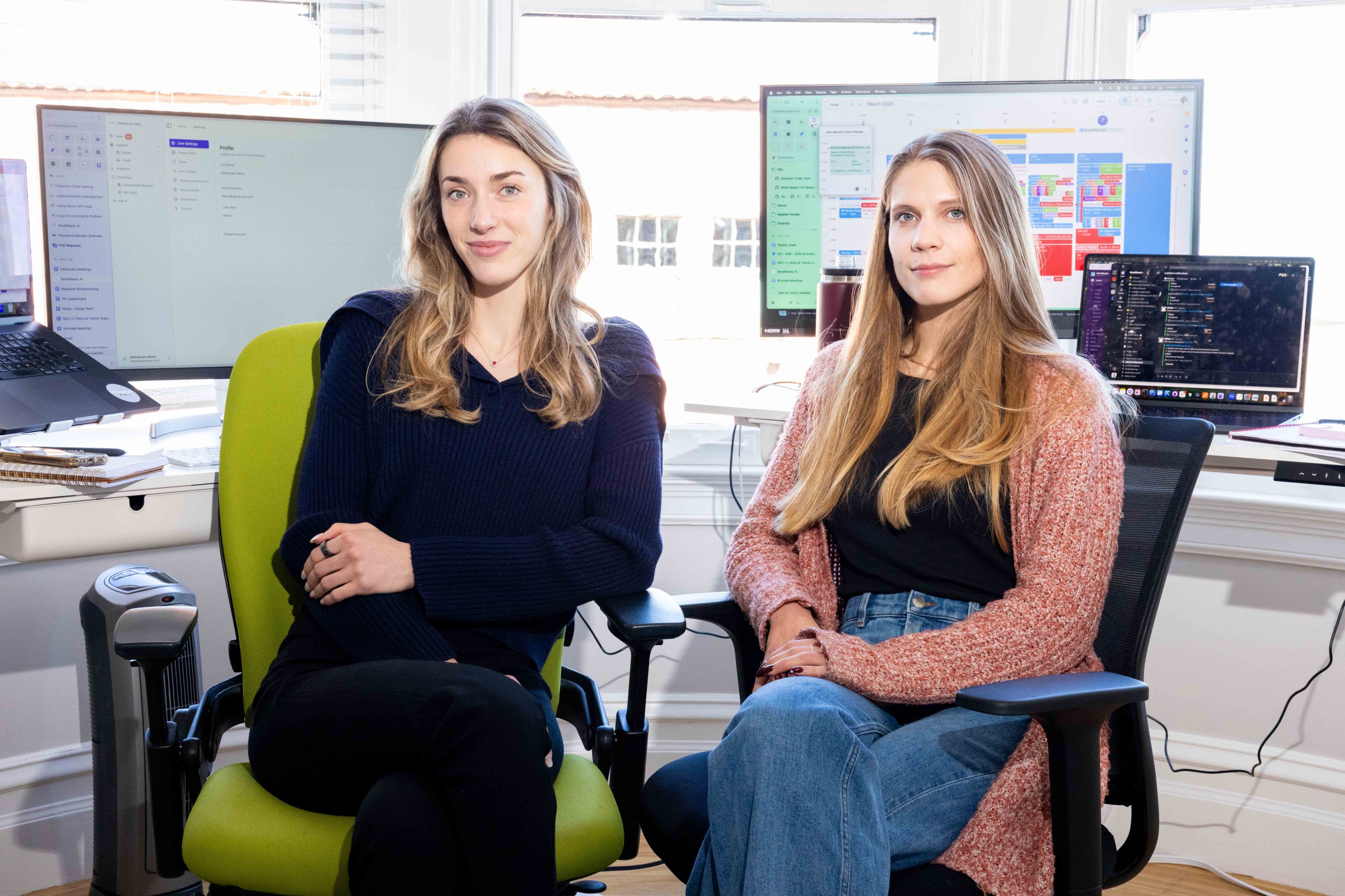 Two women are seated in an office environment. They are sitting in chairs with computer screens and laptops displaying calendars and messages in the background.