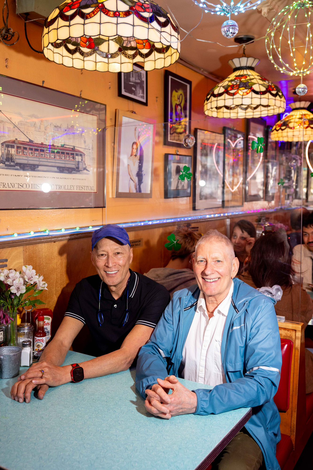 Two men sit smiling at a turquoise table in a cozy diner with vintage-style lamps and framed pictures on the walls. The atmosphere is warm and inviting.