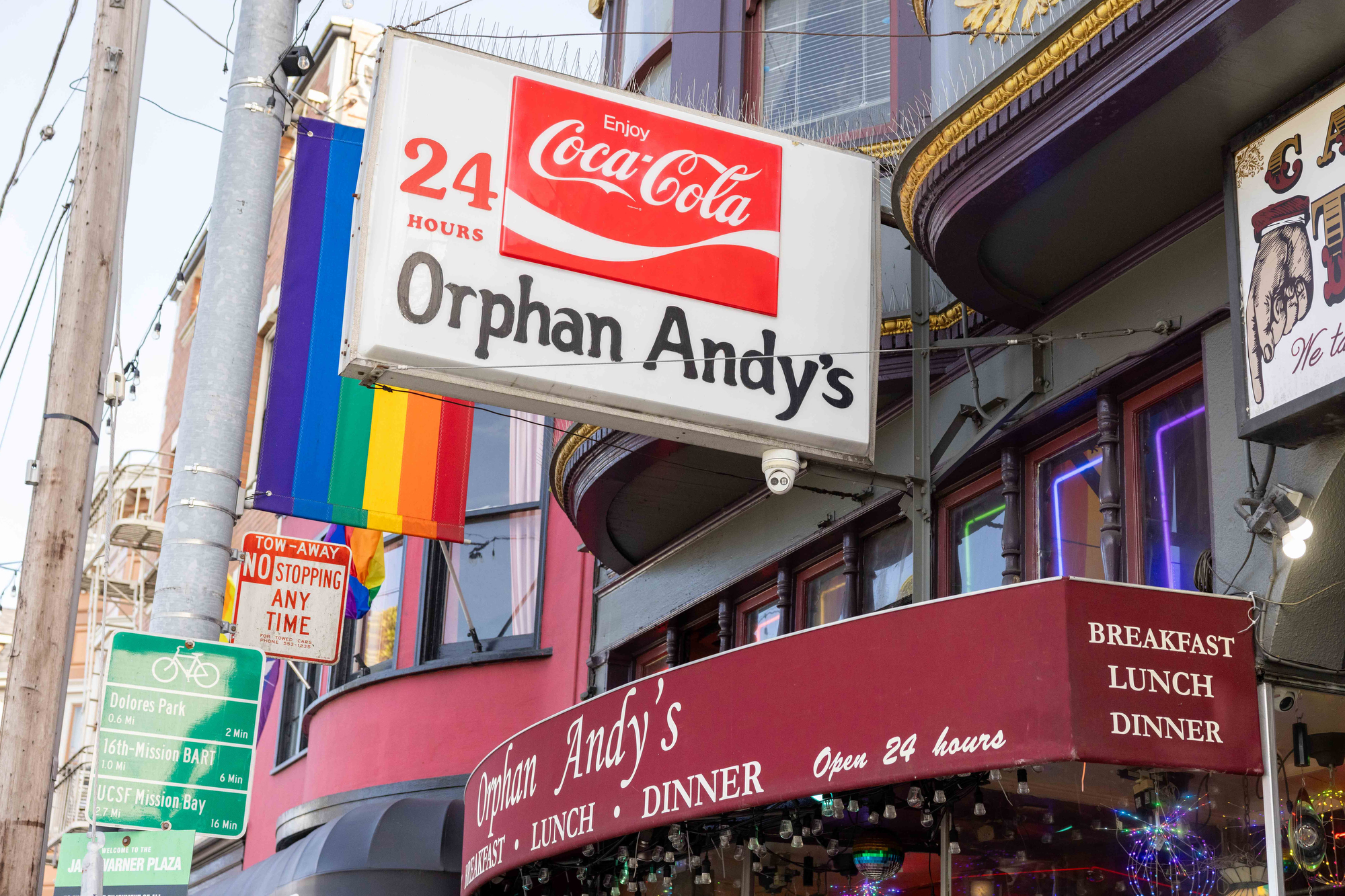 The image shows the entrance to "Orphan Andy's," a 24-hour diner. A rainbow flag is displayed prominently. Signs indicate it's open for breakfast, lunch, and dinner.