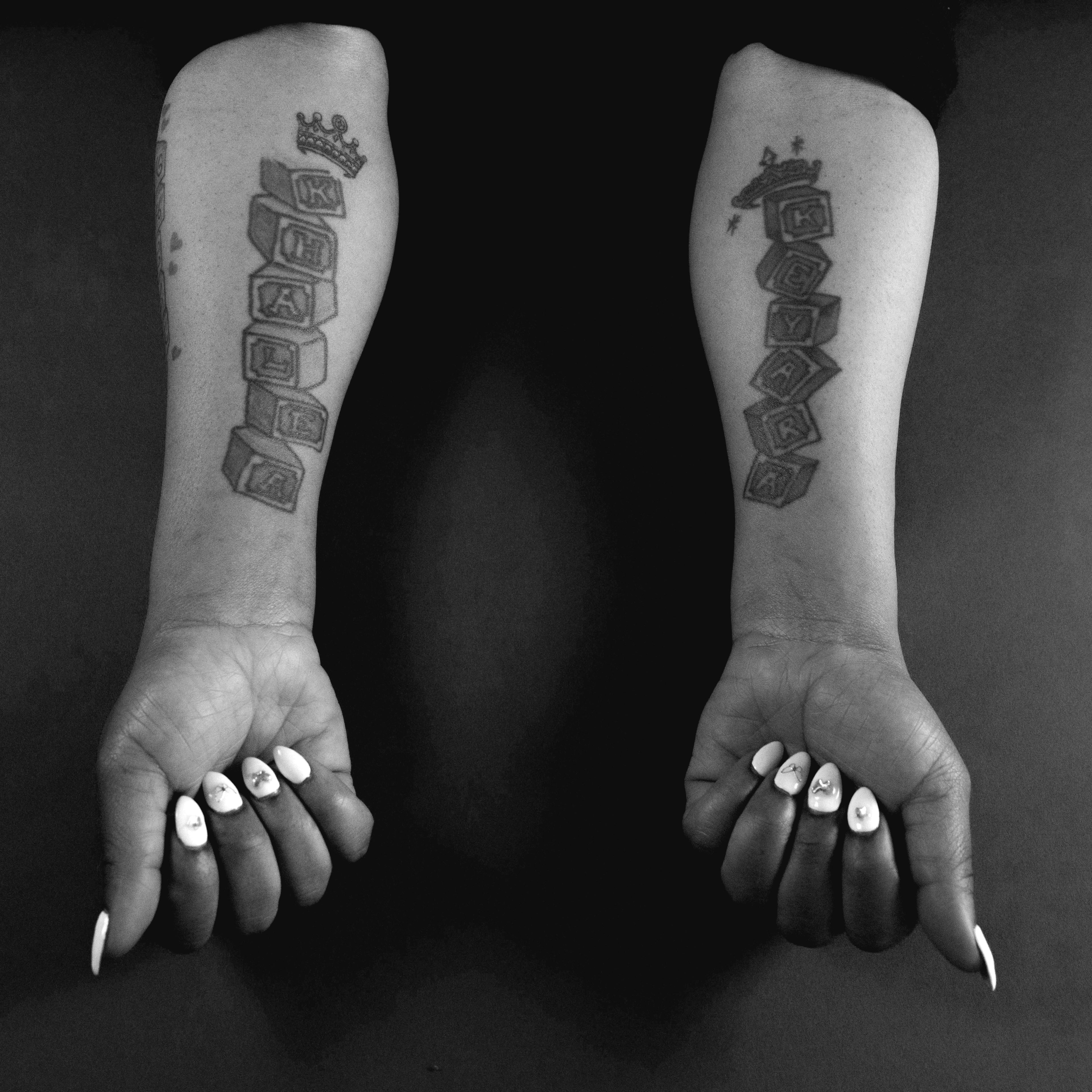 Two arms are shown with tattoos of stacked blocks spelling &quot;KHALEA&quot; and &quot;KEYARA.&quot; The nails are white with small designs.