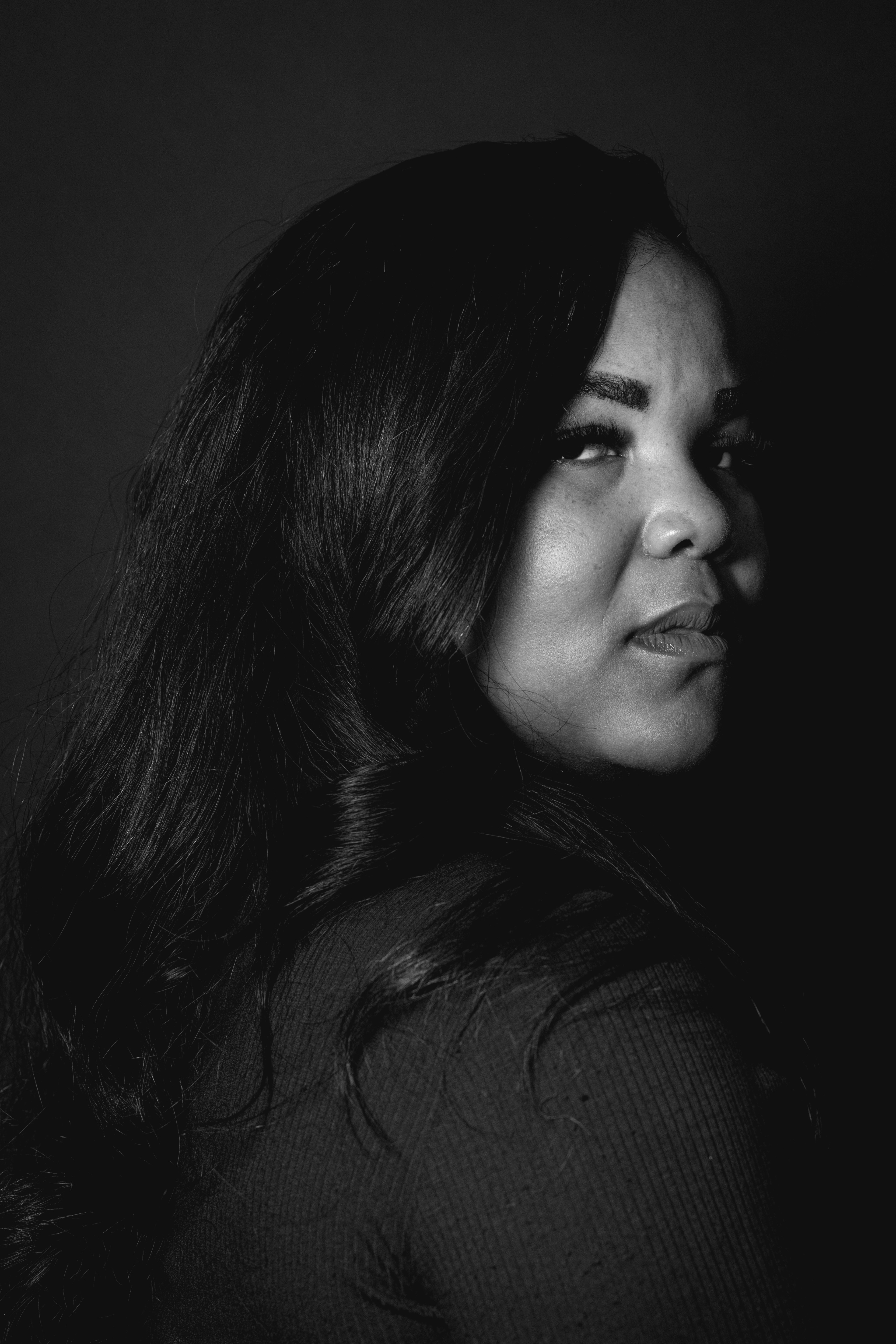 A woman with long, dark hair glances over her shoulder against a dark background. The image is in black and white, highlighting shadows on her face.