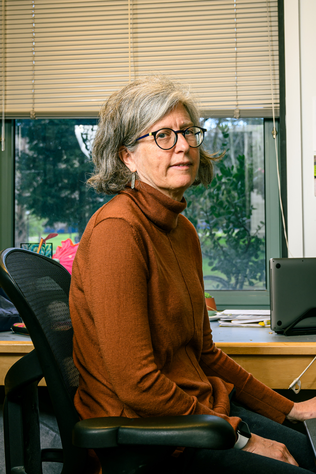 A person with gray hair, wearing glasses and a brown top, sits in an office chair. Behind them are papers and a window with blinds.