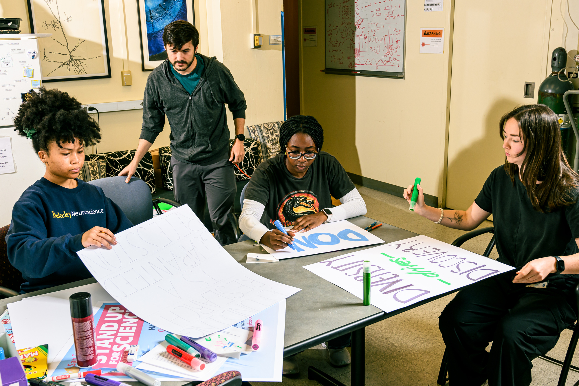 Four people are making protest signs at a table in a room with scientific posters and art on the wall. They're using markers and various supplies.