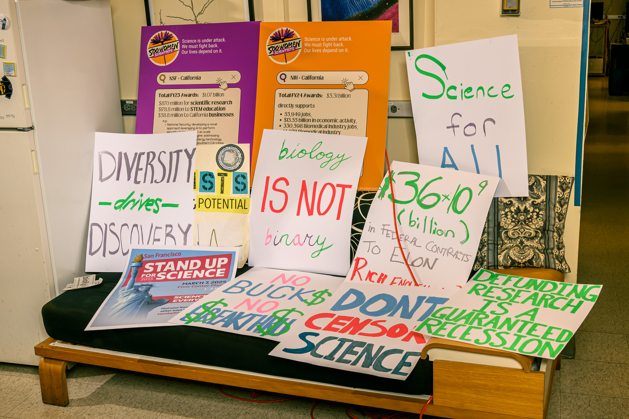 The image shows various protest signs advocating for science, diversity, and opposing censorship. Messages include &quot;Diversity drives discovery&quot; and &quot;Biology is not binary.&quot;