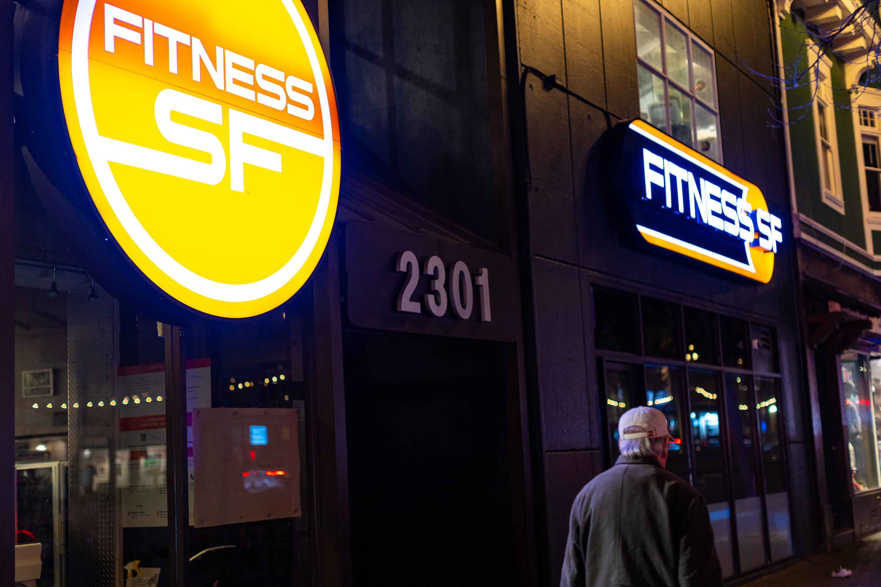 A person in a cap walks on a nighttime street past a brightly lit &quot;Fitness SF&quot; sign with the address &quot;2301&quot; visible.