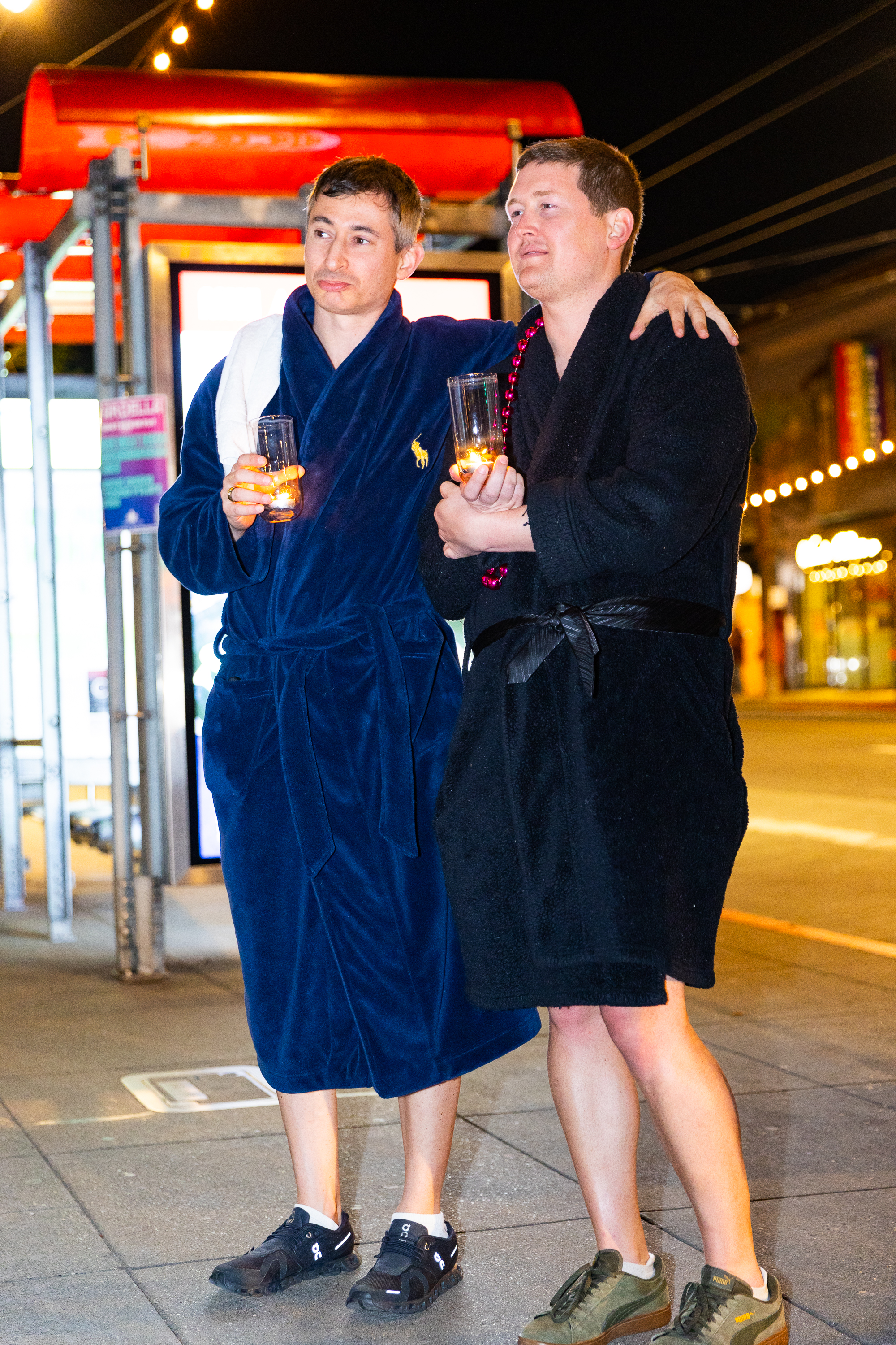 Two people in robes stand on a street at night, hugging and holding glasses with candles. One wears a blue robe, the other a black robe.