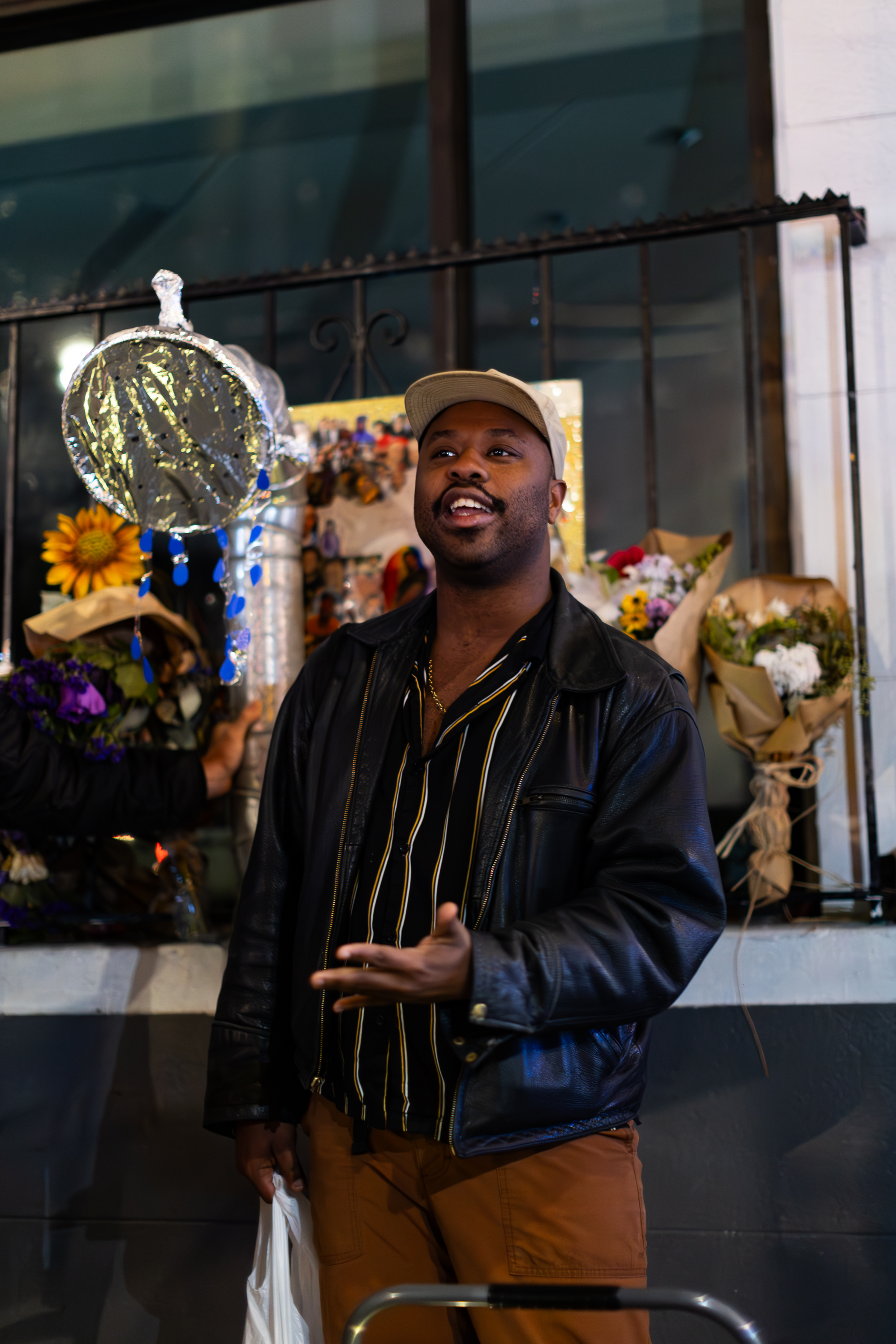 A person in a leather jacket and cap gestures while holding a bag. Behind them are flowers and a silver decoration hanging nearby.