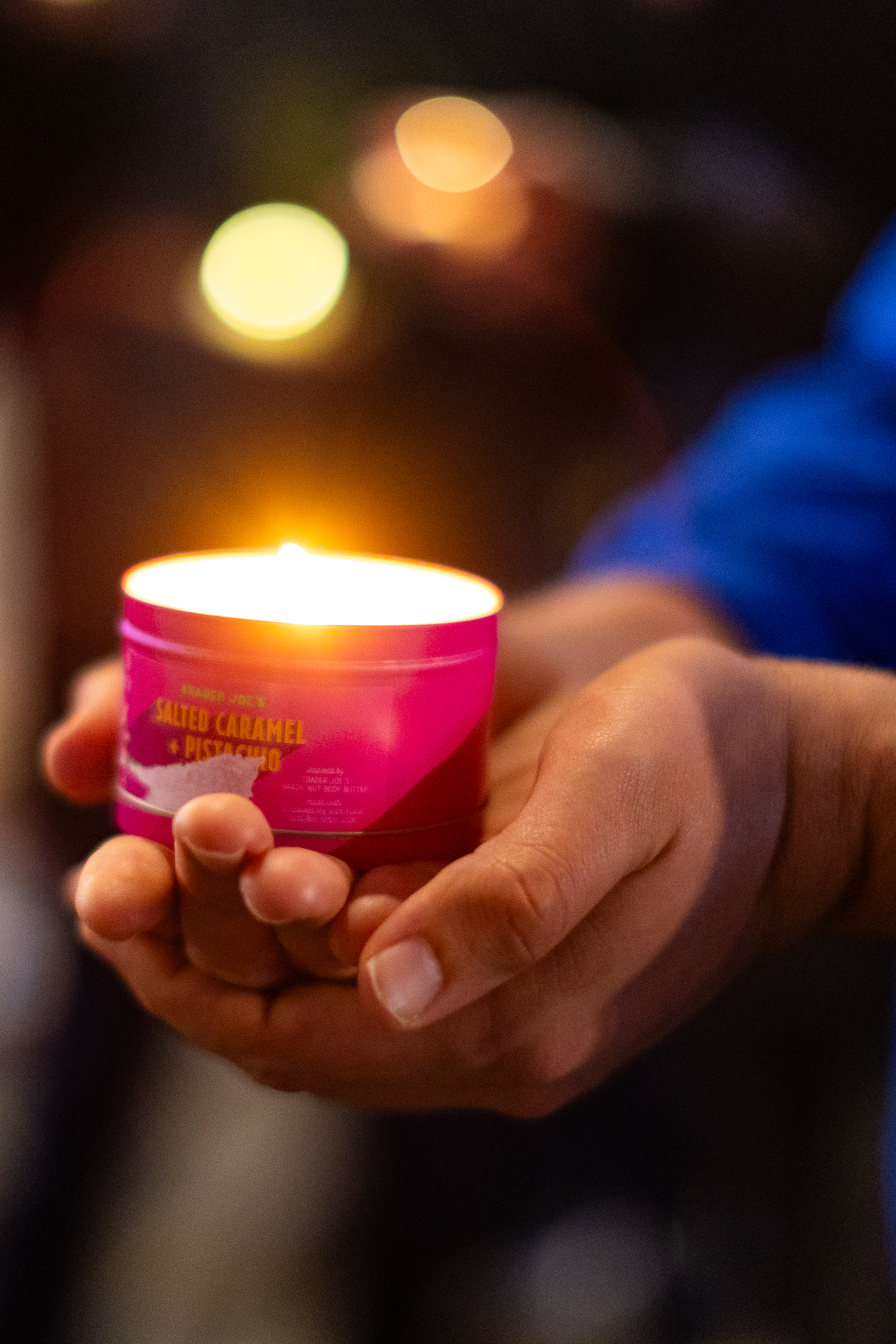 A person is holding a lit candle with a pink label in their cupped hands. The label reads &quot;Salted Caramel and Pistachio.&quot; The background is softly blurred.