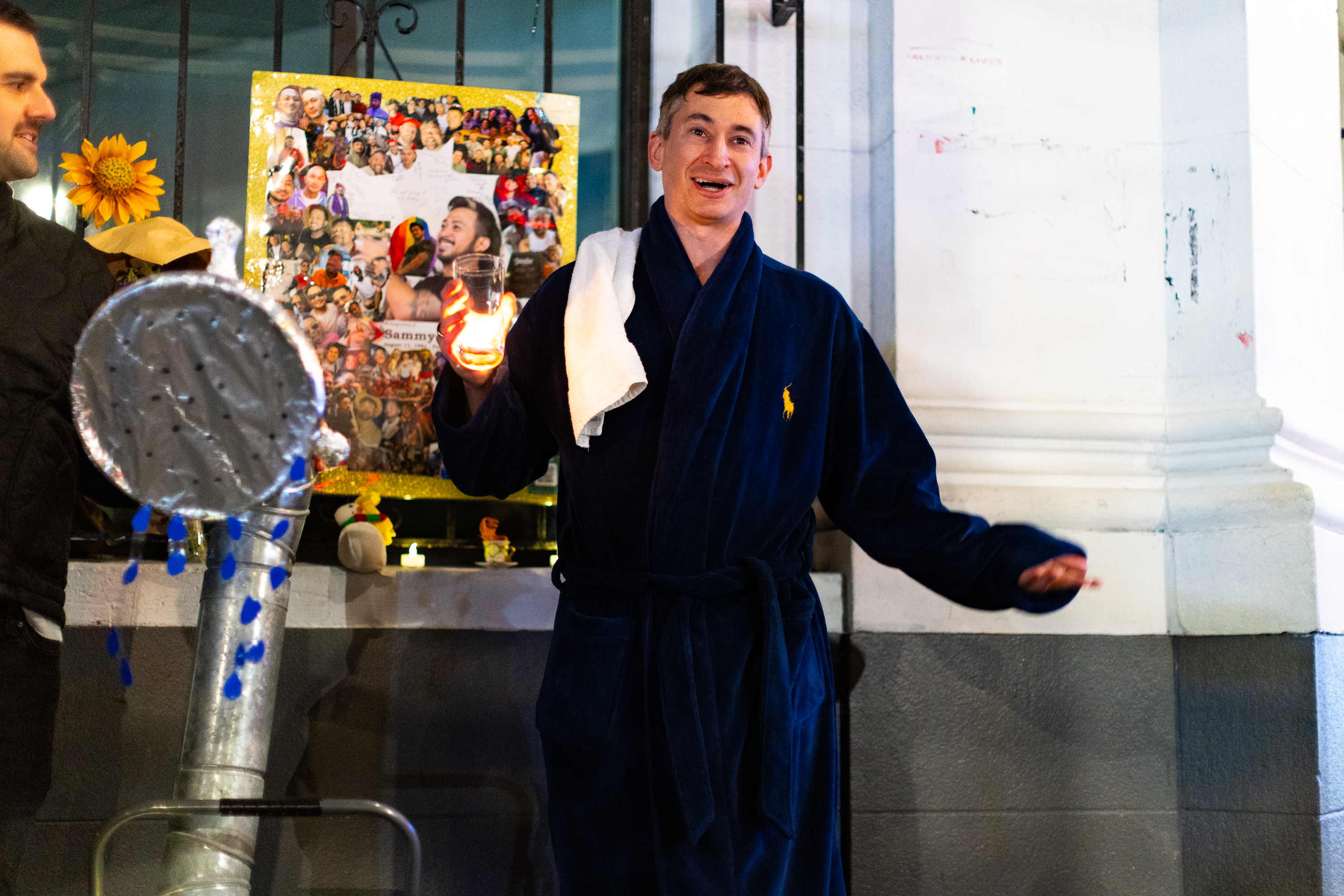 A person wearing a dark robe and holding a jar with a light stands in front of a collage of photos. A large silver prop is in the foreground.