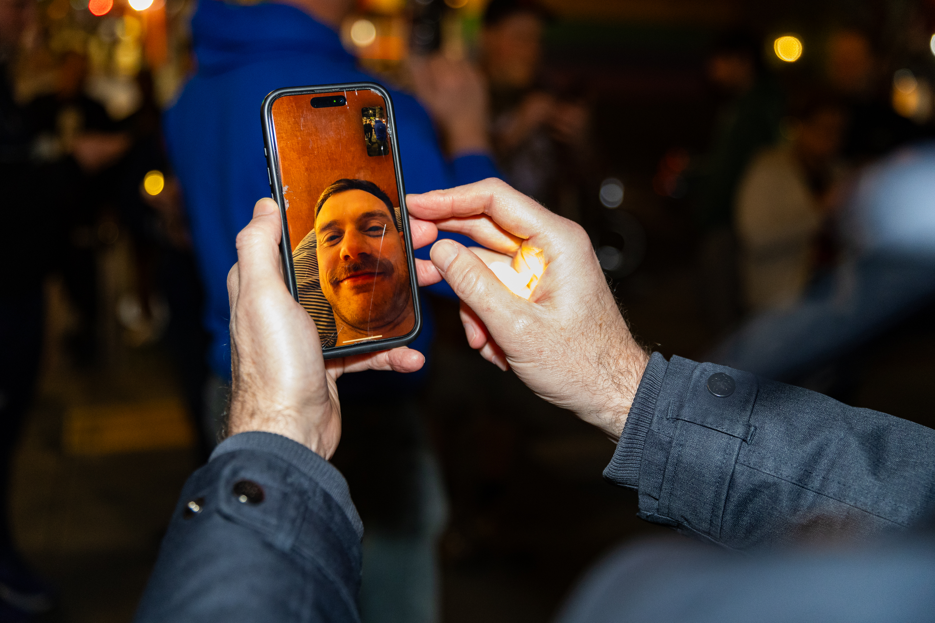 A person holds a phone showing a video call with someone smiling. The call is happening at night, and the person is using their other hand to shield the screen from light.