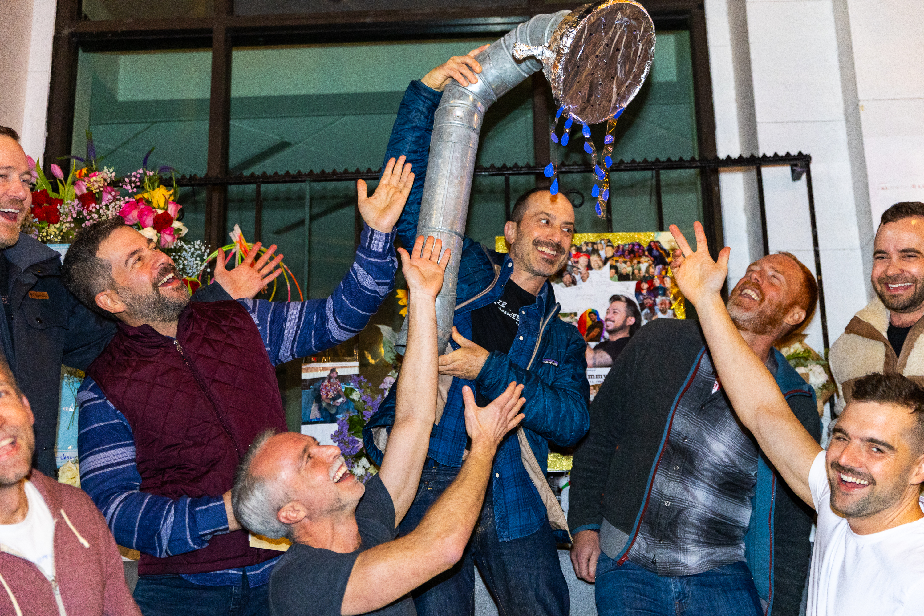 A group of smiling men gather around a metallic pipe sculpture with decorations, raising their hands joyfully. There are colorful flowers and photographs in the background.