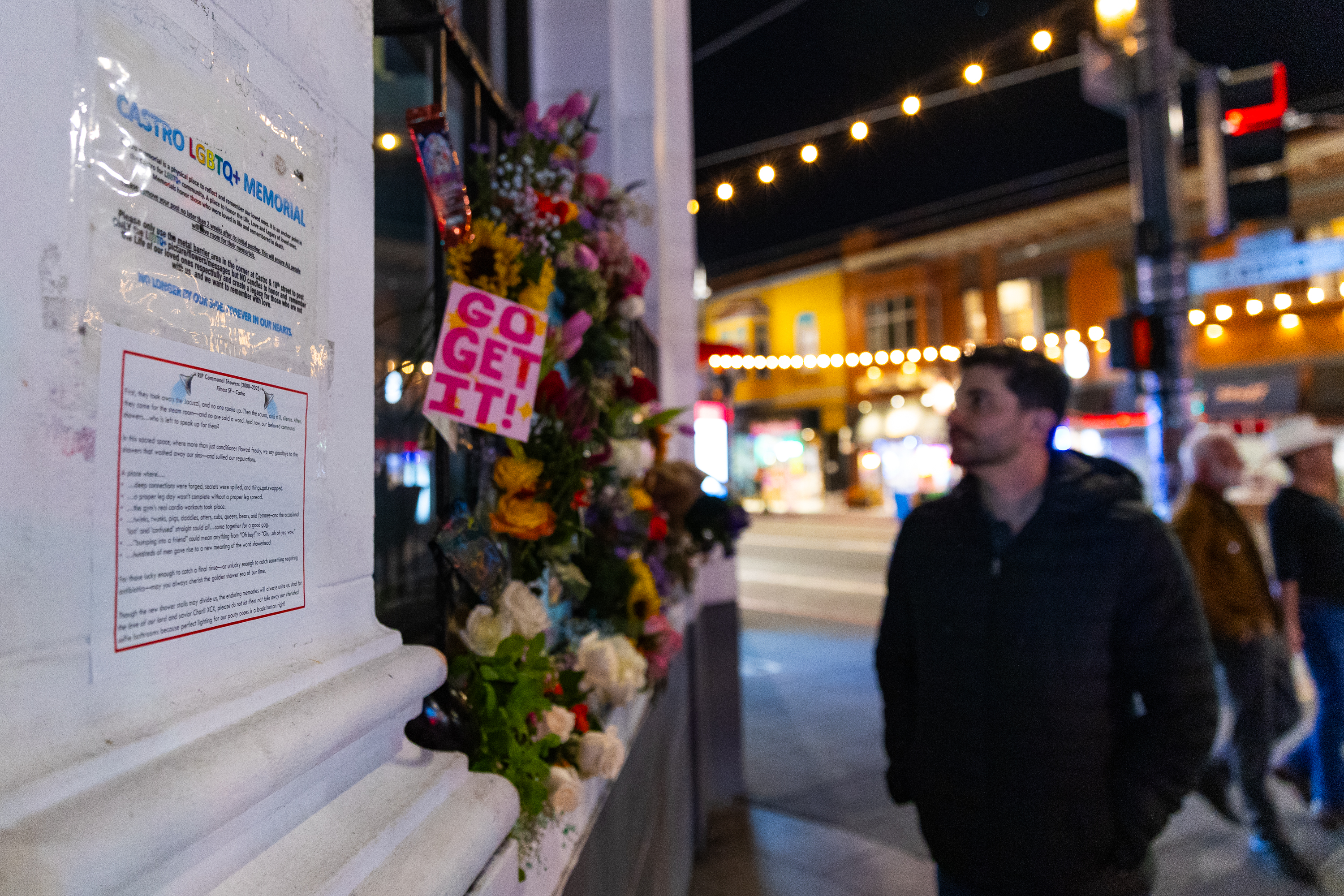 A man stands on a lit street at night, observing a memorial with flowers and a "Go Get It!" sign. Nearby, there's a sign titled "Castro LGBTQ+ Memorial."