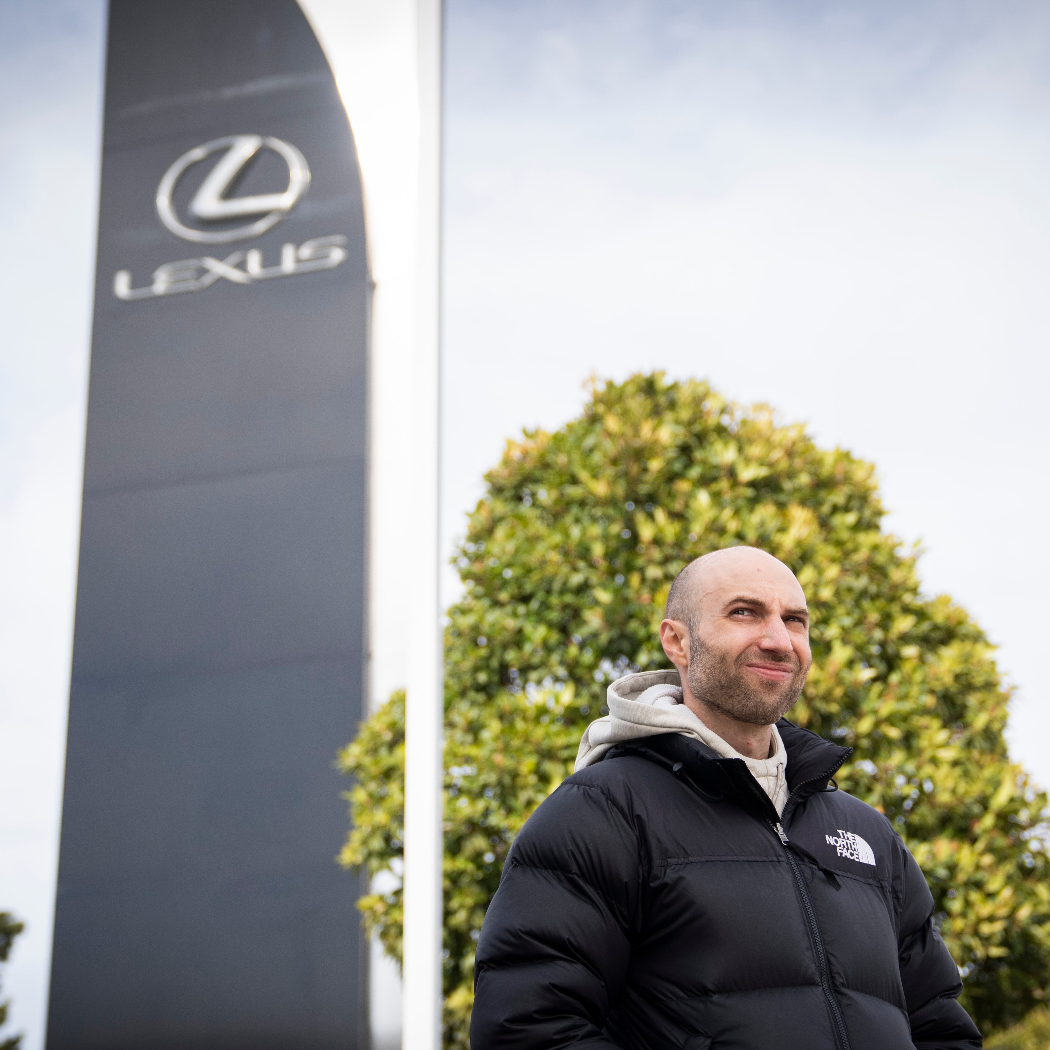 A person wearing a black puffer jacket stands outdoors near a large sign with the Lexus logo, against a backdrop of trees and a cloudy sky.
