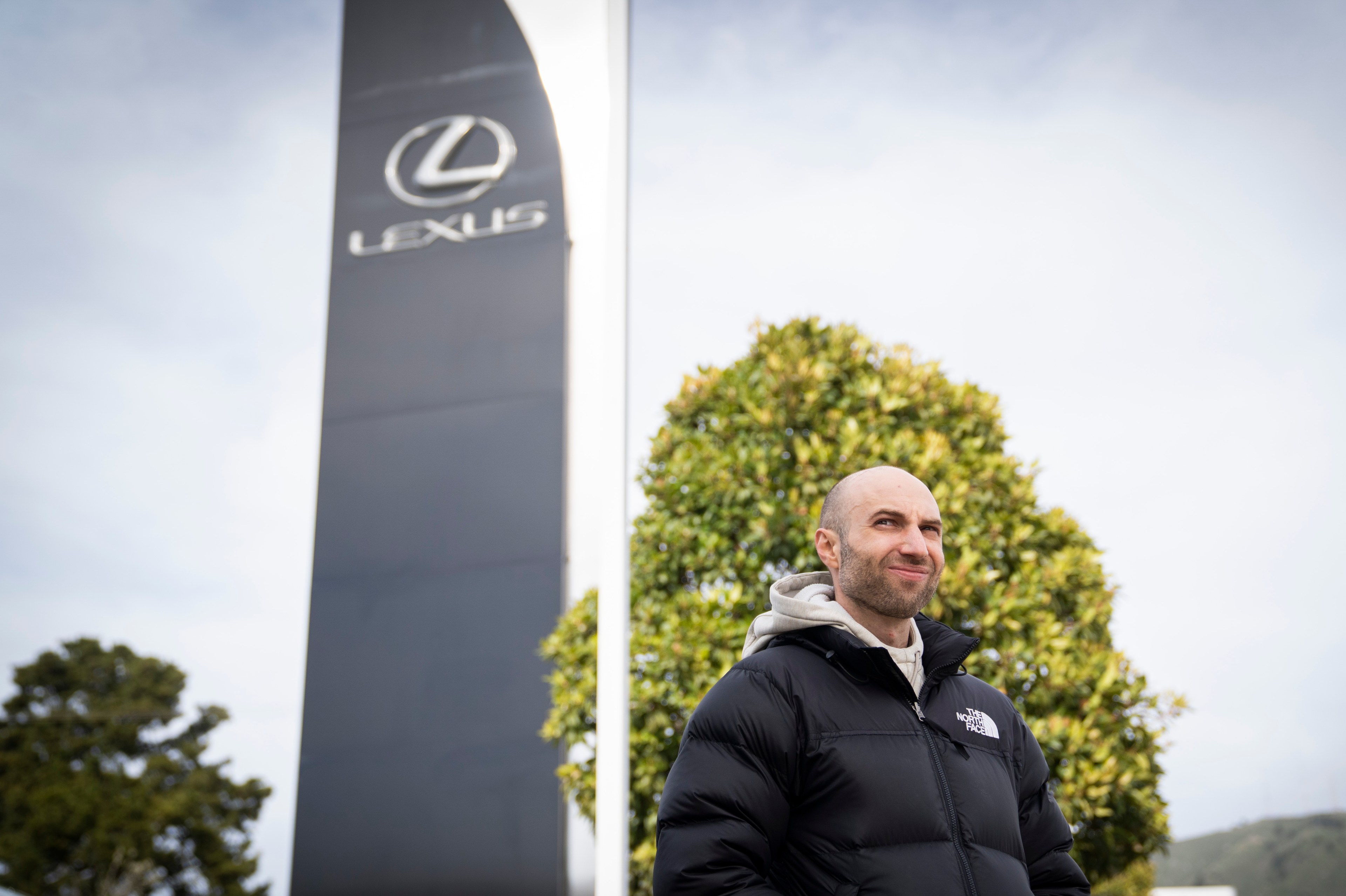 A person wearing a black puffer jacket stands outdoors near a large sign with the Lexus logo, against a backdrop of trees and a cloudy sky.