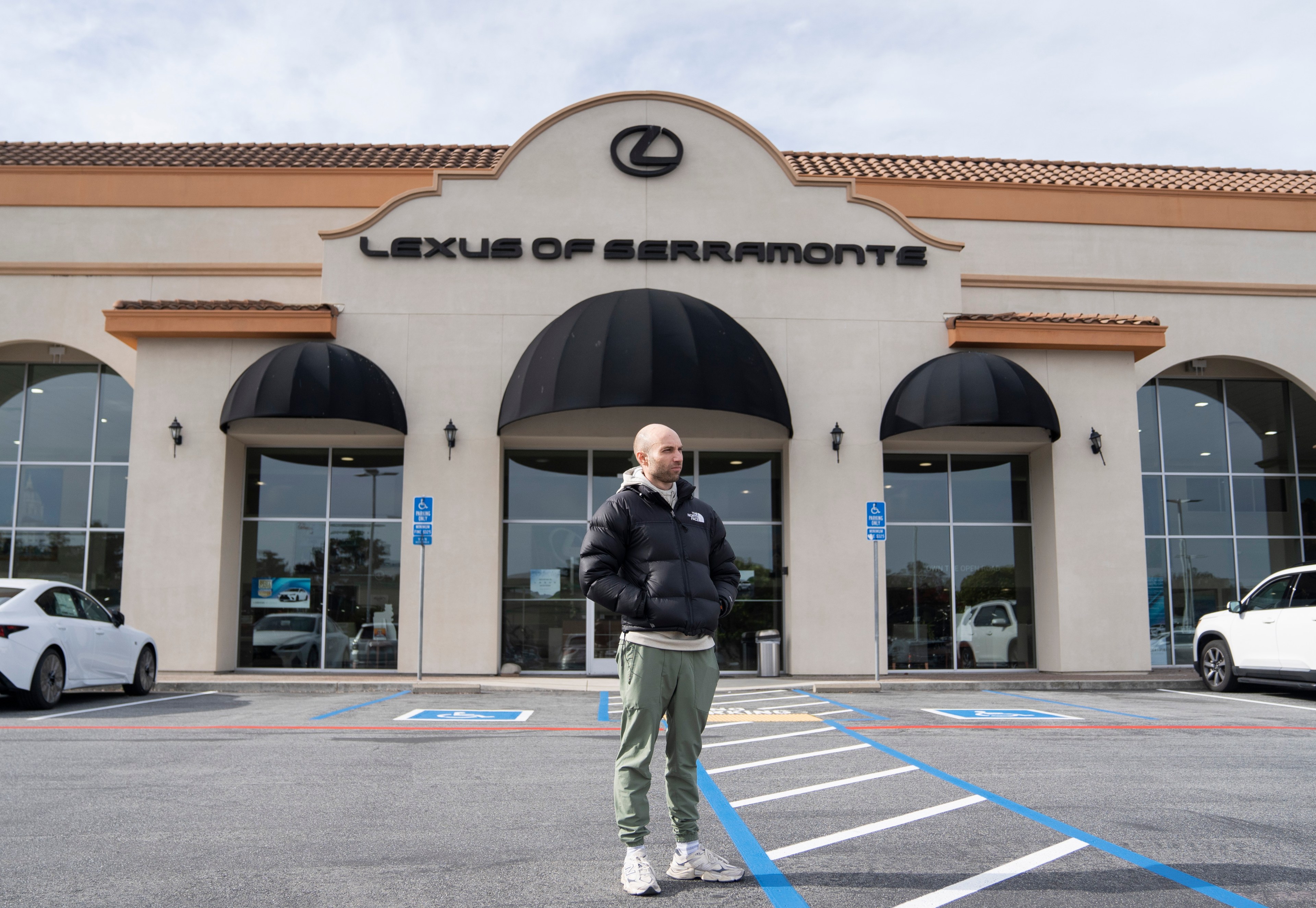 A person in a black puffer jacket and green pants stands outside a &quot;Lexus of Serramonte&quot; dealership with large windows and cars parked nearby.