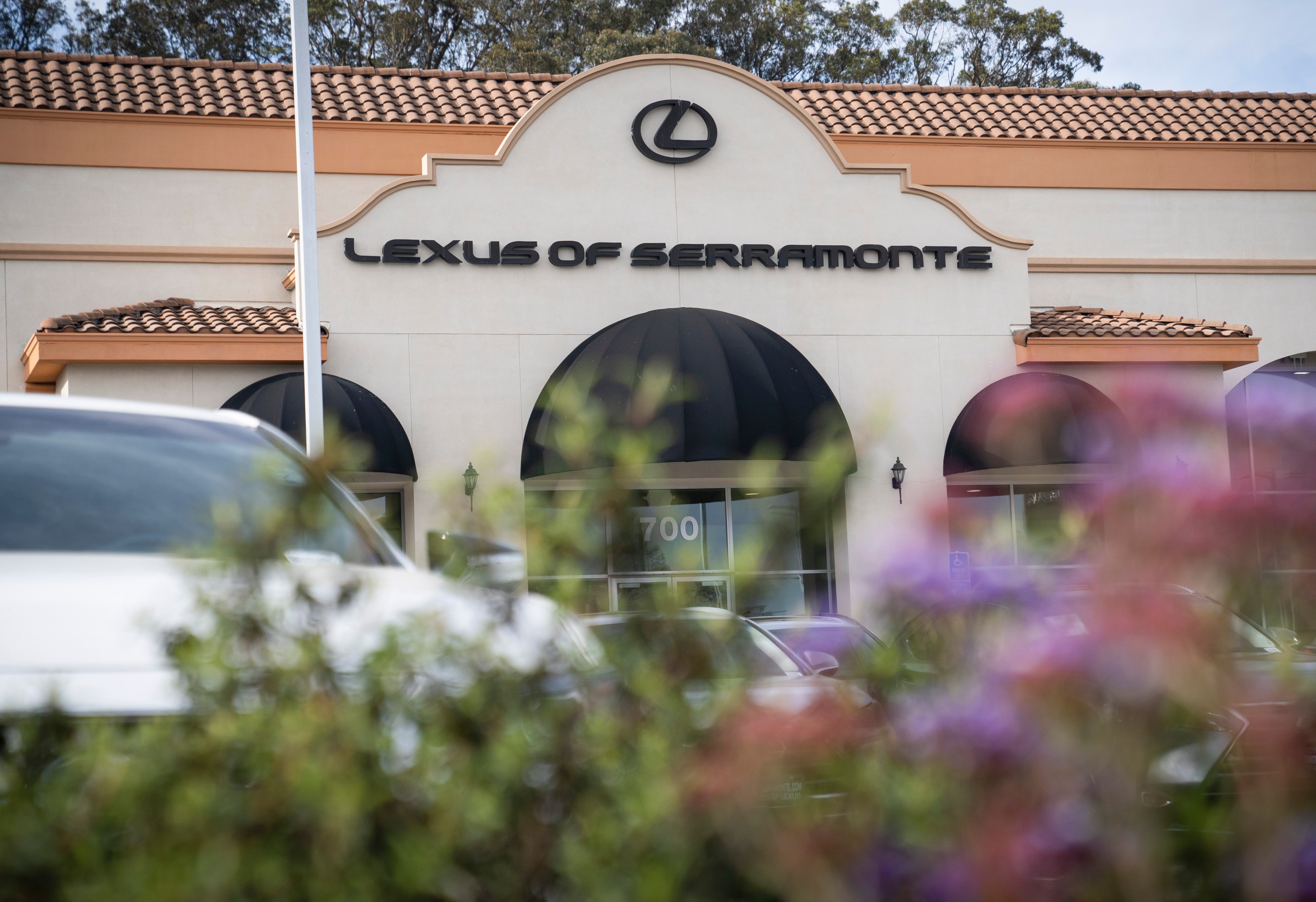The image shows the entrance of a Lexus dealership, &quot;Lexus of Serramonte,&quot; with a clay-tiled roof and black awnings, partially obscured by plants.