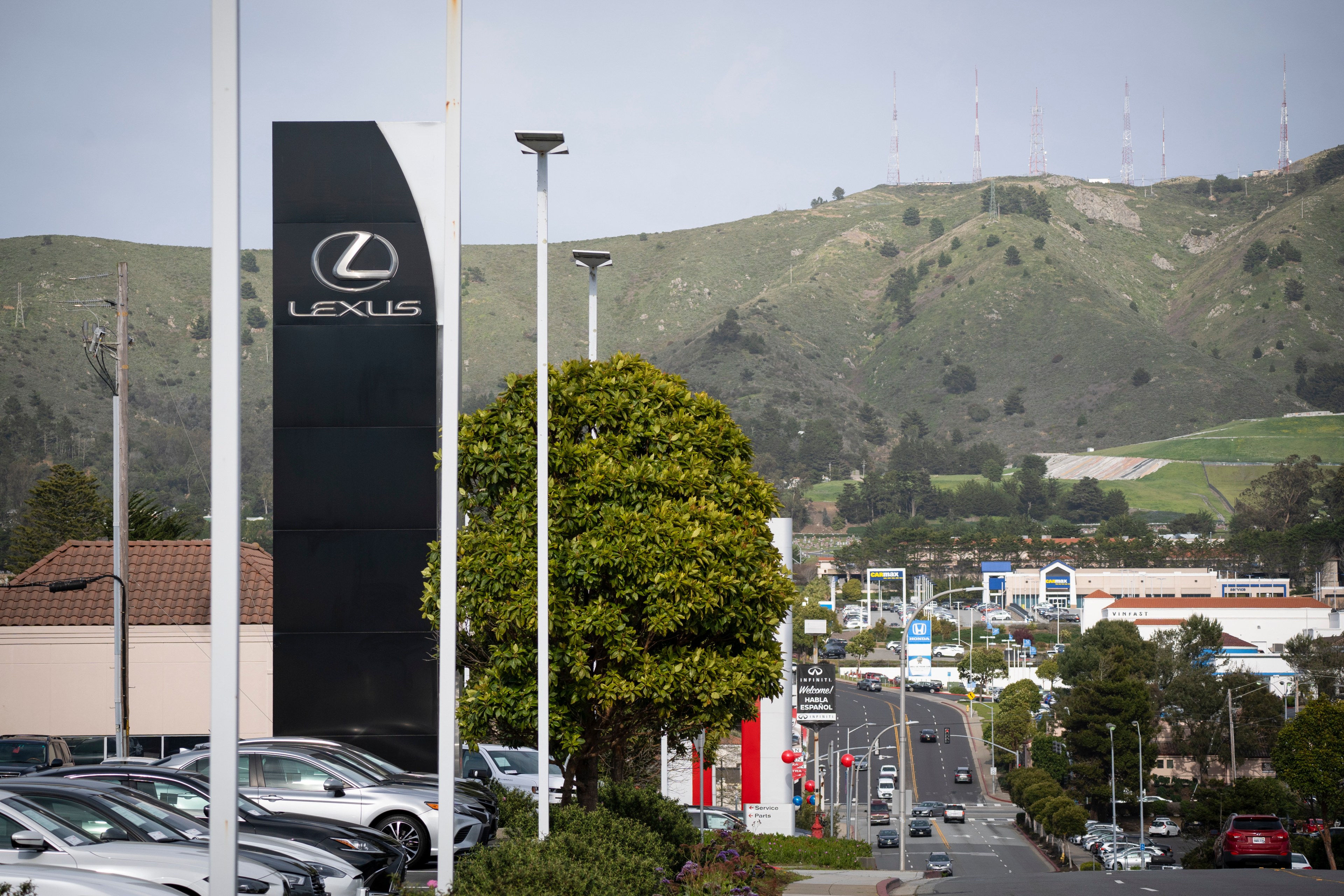 A Lexus dealership sign stands beside parked cars, with a tree and streetlights nearby. In the background, a road leads toward hills and transmission towers.