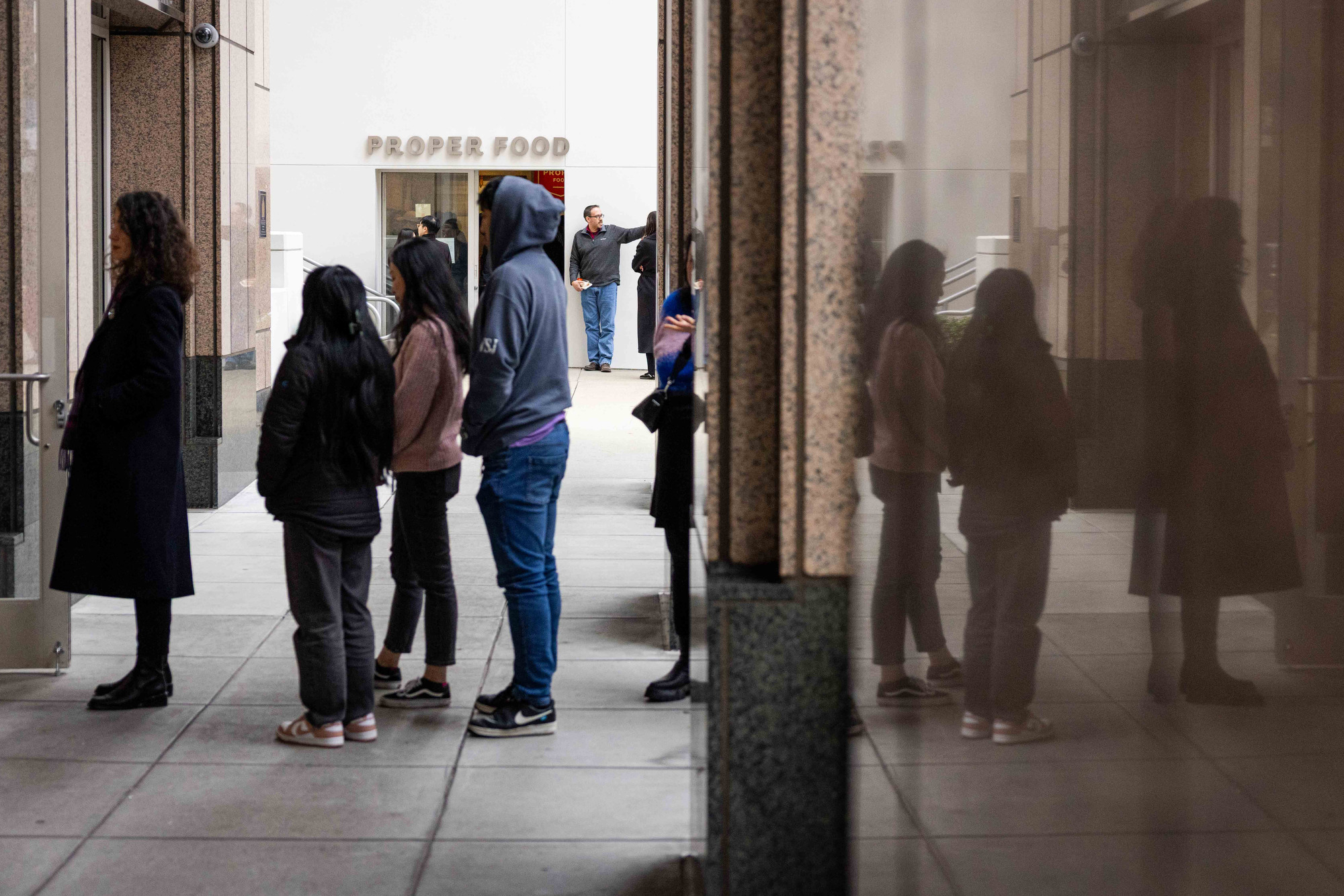 A line of people wait outside a building with &quot;Proper Food&quot; written above the entrance. Their reflection is visible on the adjacent glass wall.