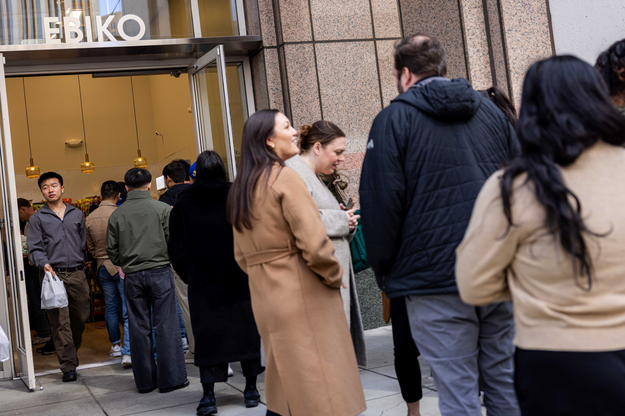 A group of people is waiting in line outside a store named &quot;EBIKO.&quot; The entrance is open, and people are entering, with some carrying bags.