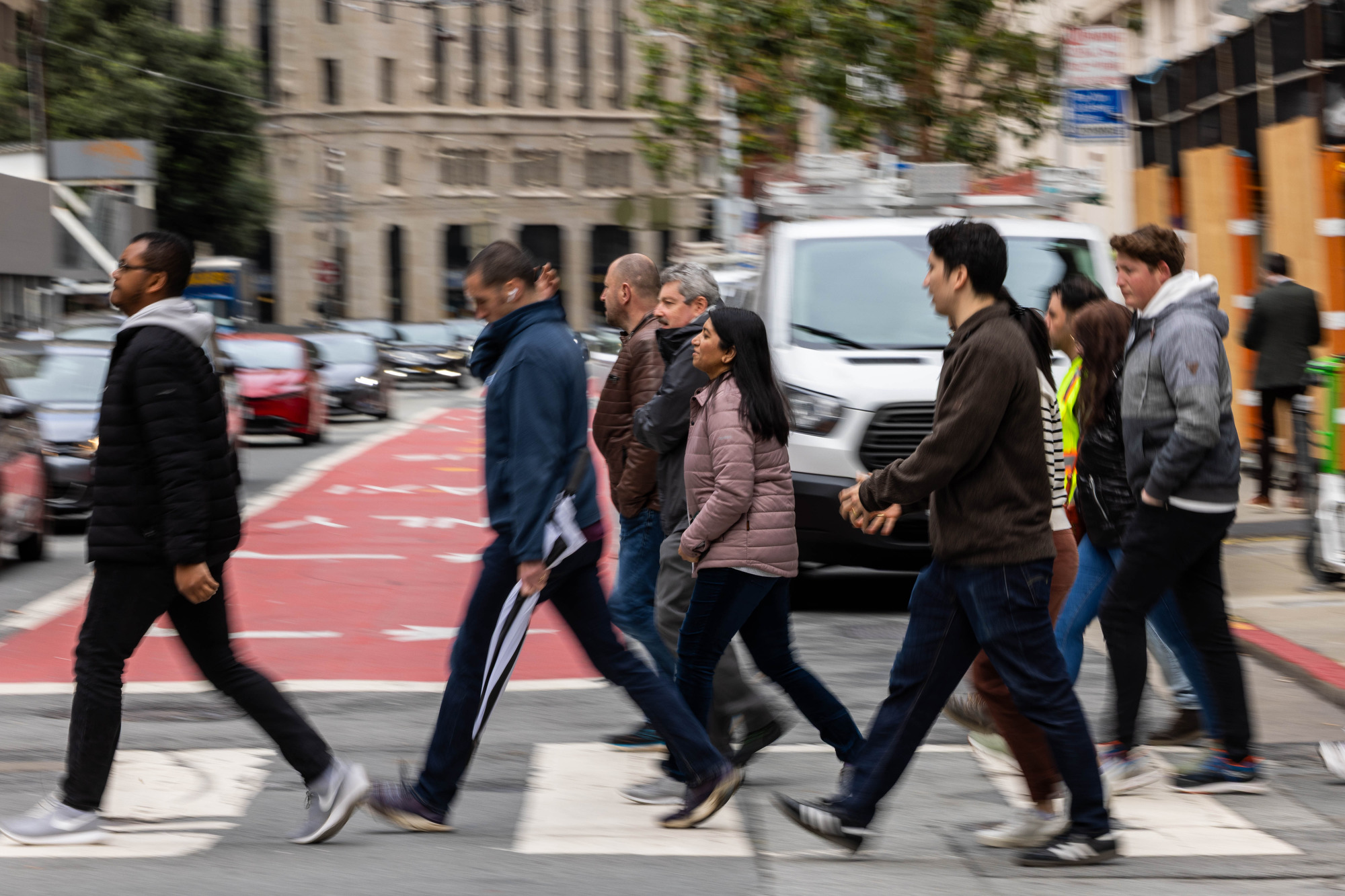 A group of people crosses a city street at a crosswalk. They walk briskly, while cars are stopped. A red bus lane and buildings are visible in the background.