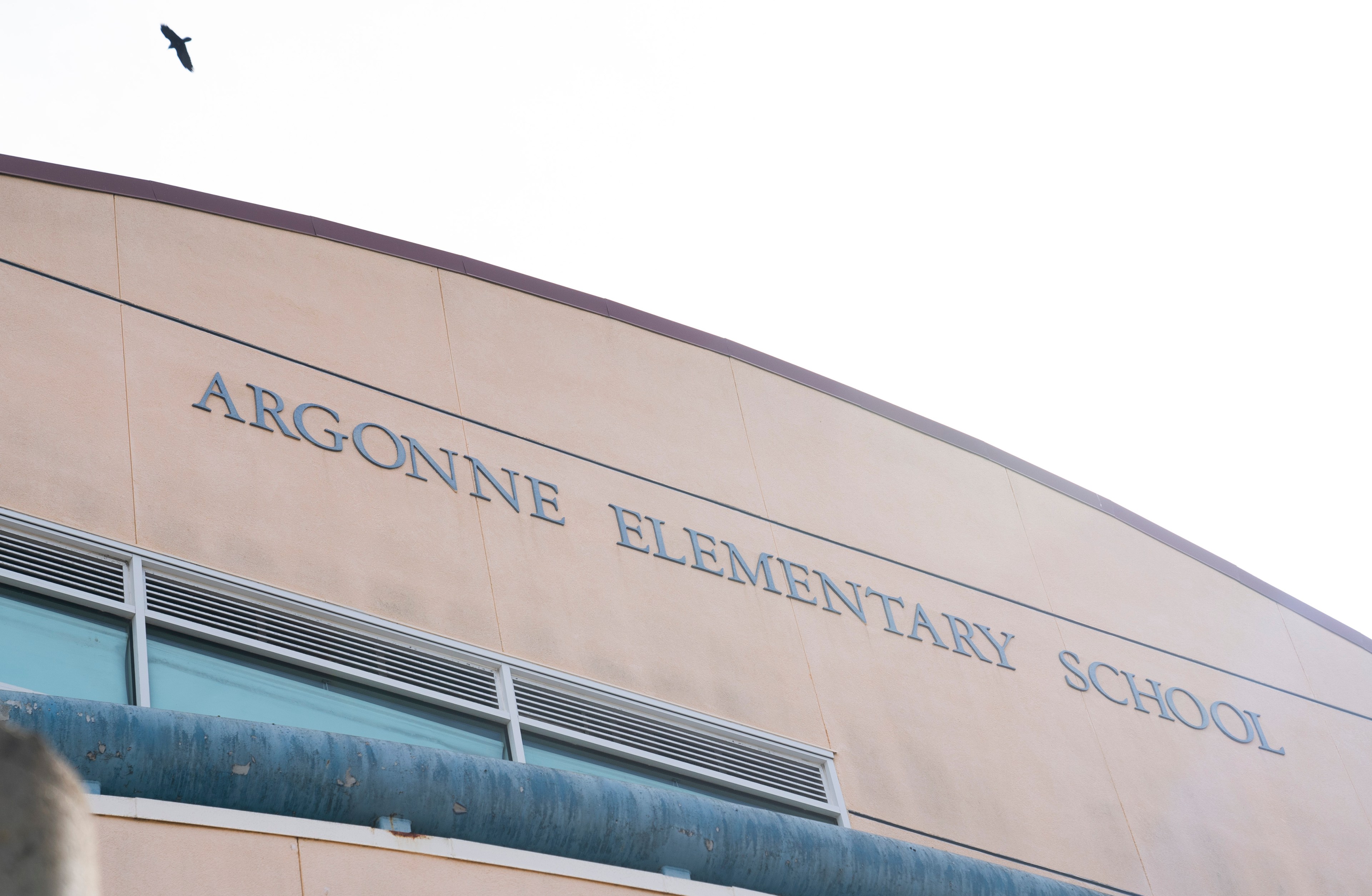 The image shows the beige facade of Argonne Elementary School with its name on the wall. A black bird flies above against a pale sky.