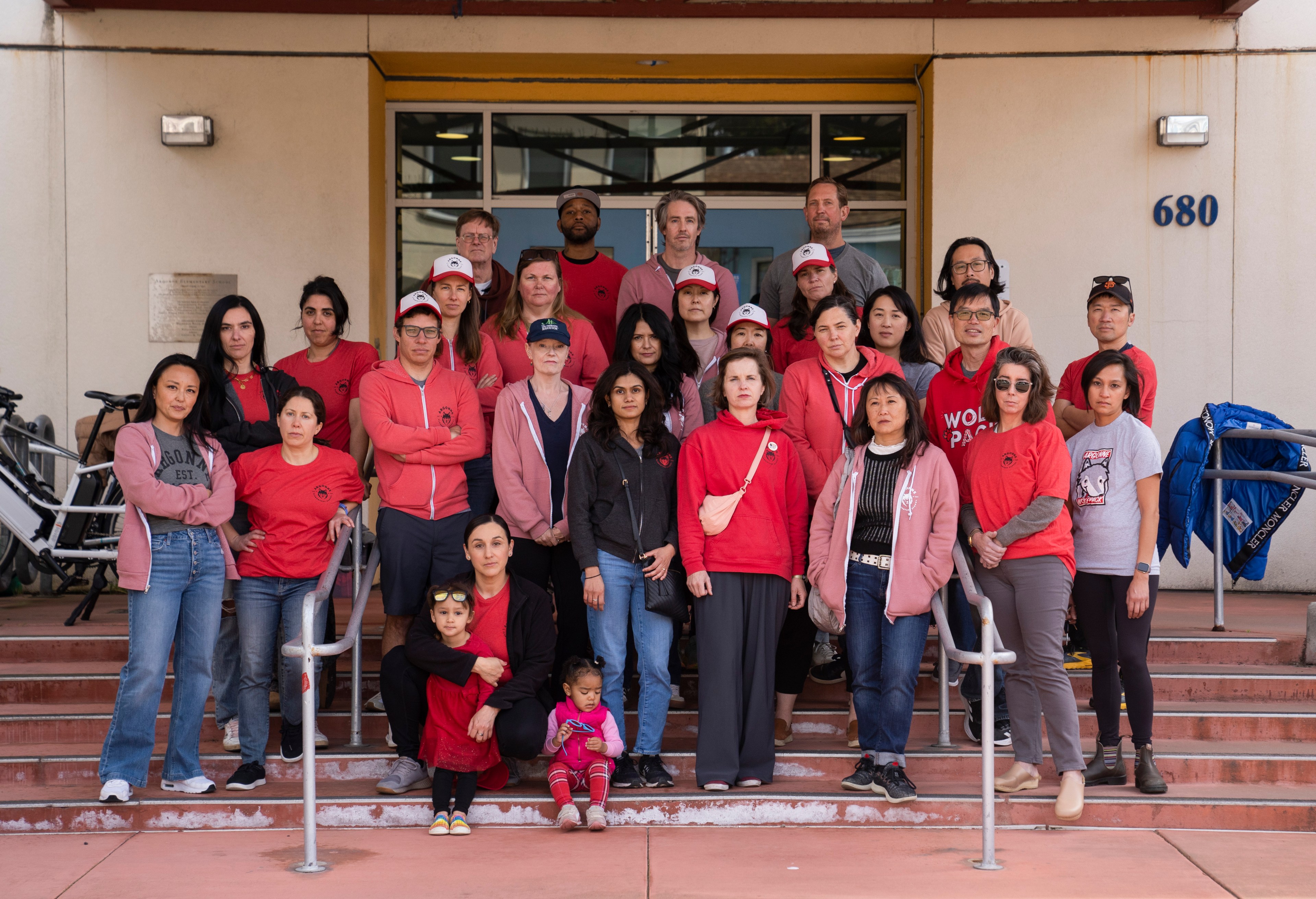 A diverse group of adults and children, some wearing red, pose on steps in front of a building numbered 680. They appear serious and united.